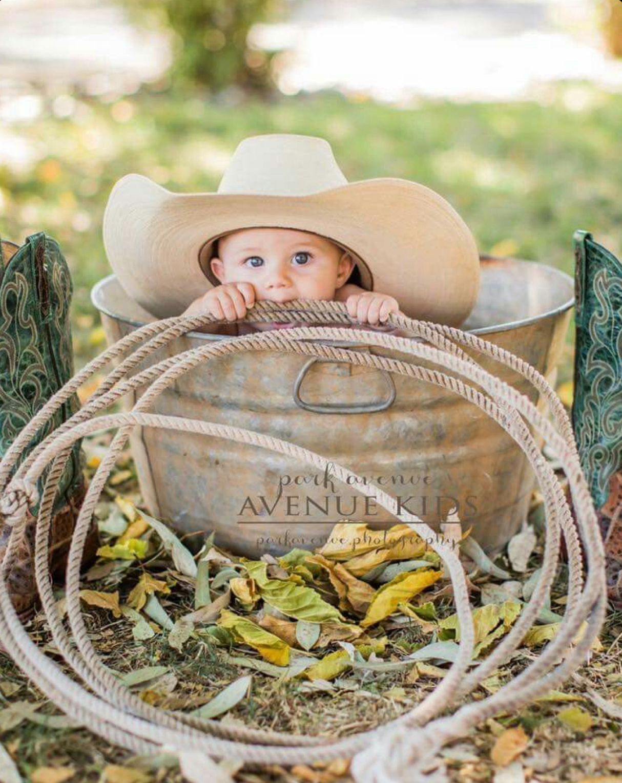 Adorable baby in a cowboy hat sitting in a rustic galvanized tub with a rope, surrounded by autumn leaves.