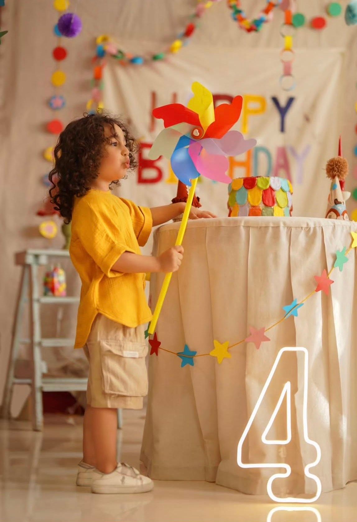 Young child in yellow shirt holds colorful pinwheel at decorated 4th birthday party with Happy Birthday banner.