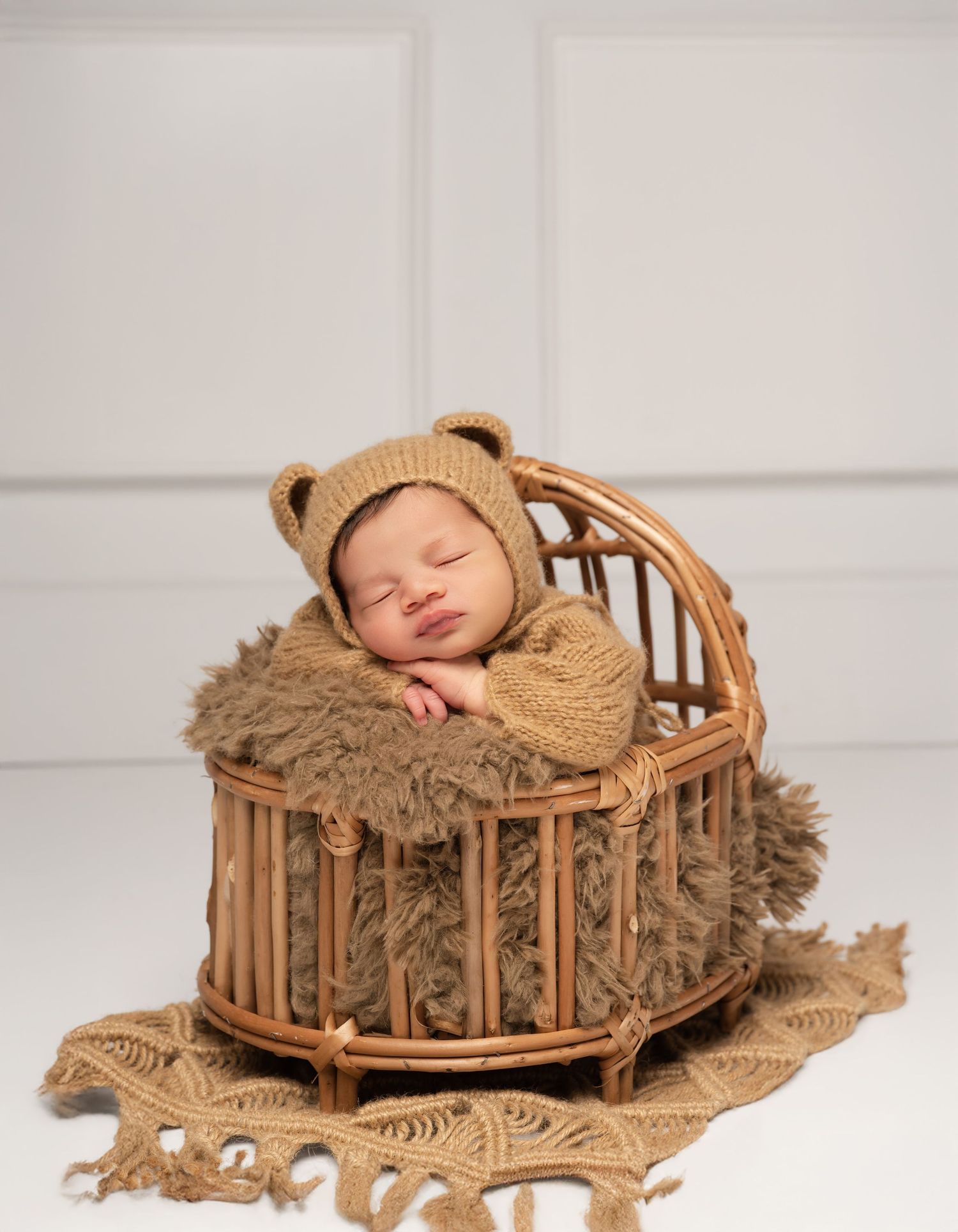 Newborn baby sleeping in a wicker basket wearing a knit bear bonnet, nestled in fluffy brown fur.
