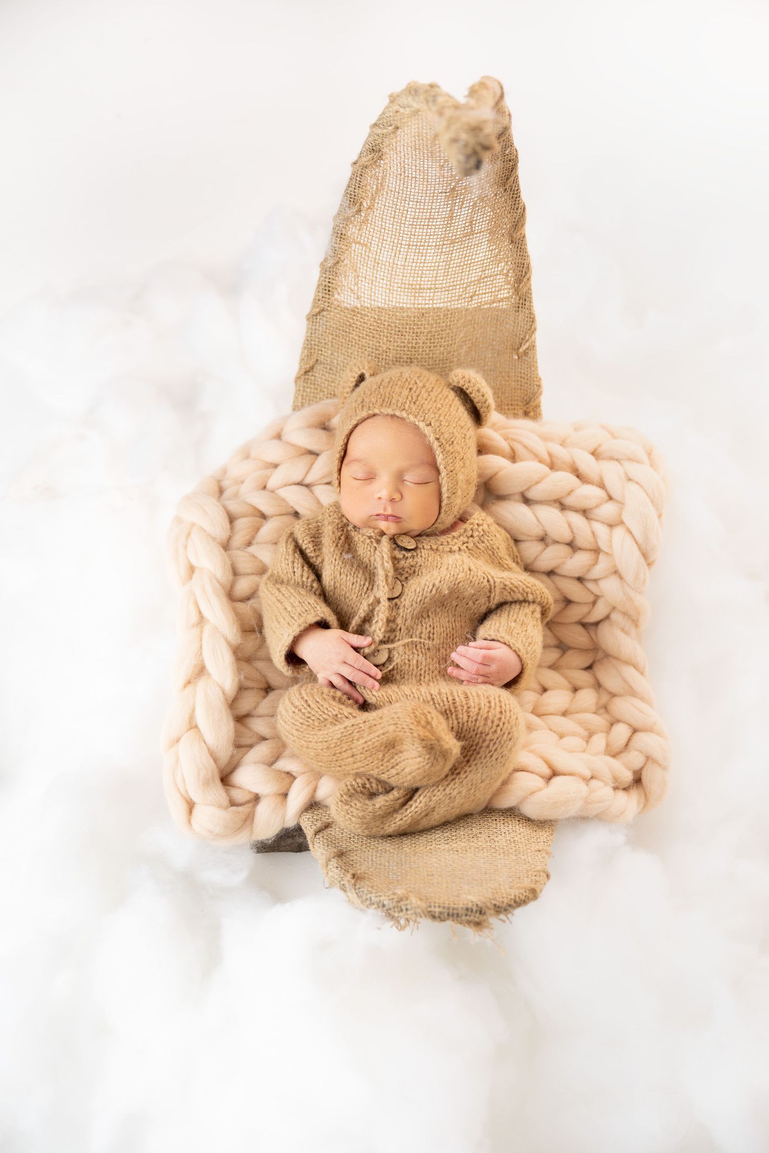 Newborn baby sleeping in bear outfit on chunky knit blanket in wooden crate with white fluffy background.
