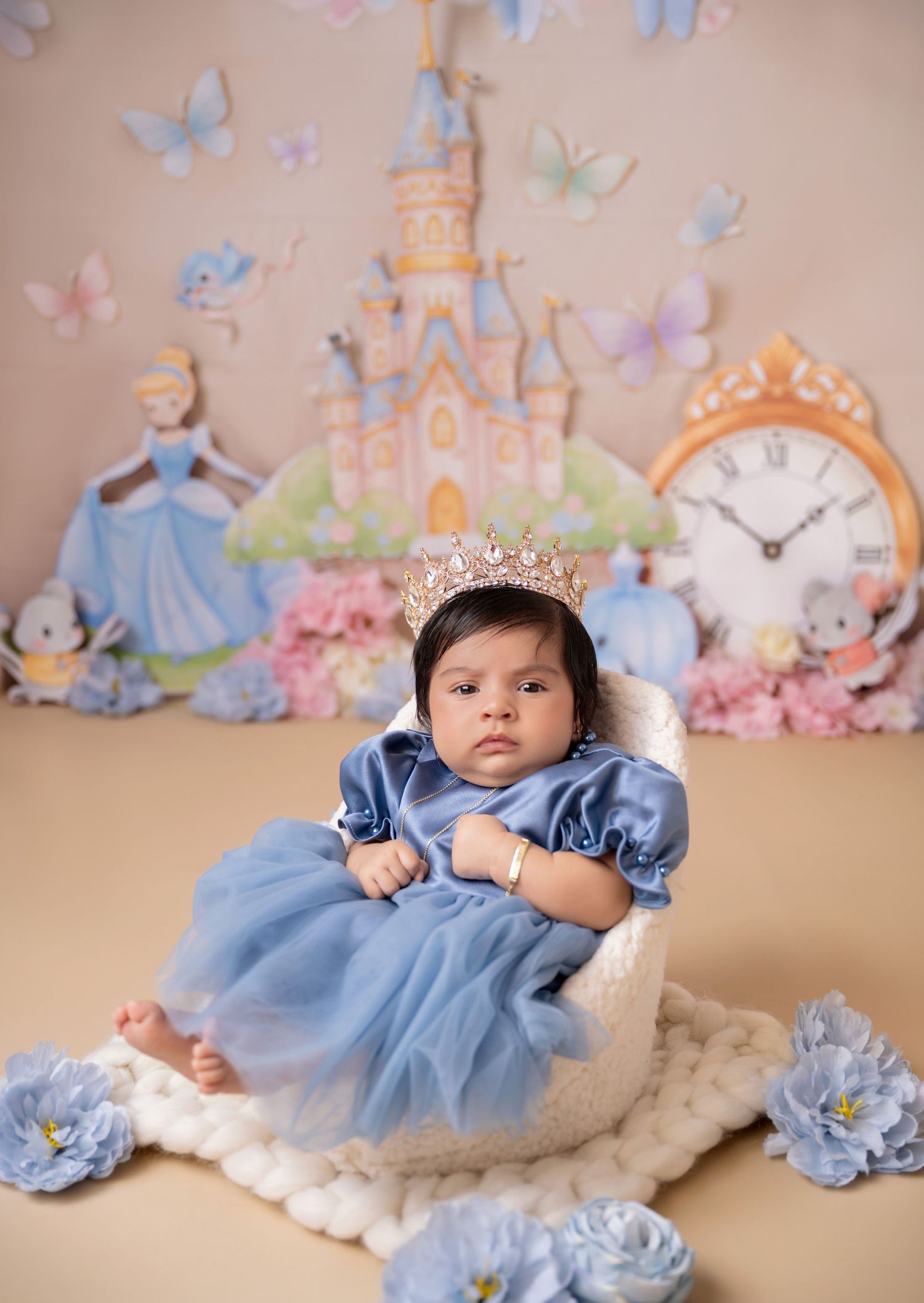 Baby girl in blue Cinderella dress and crown sits in white chair with fairytale castle backdrop and butterflies.