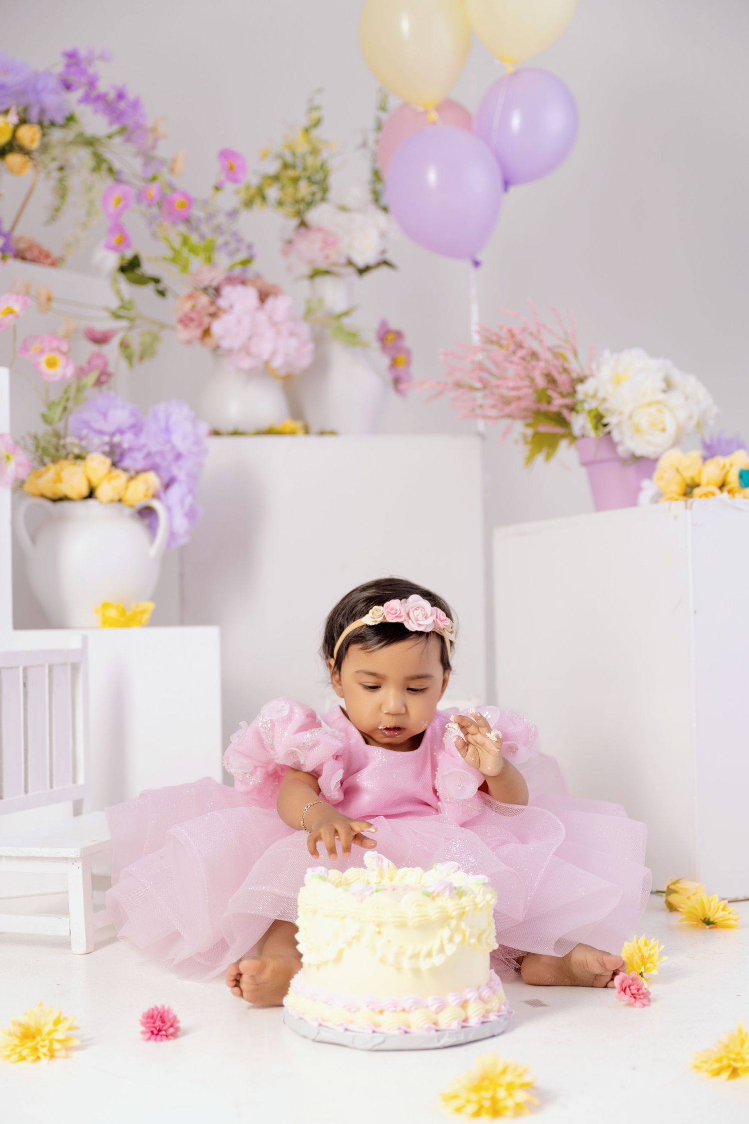 Baby girl in pink tutu dress reaching into birthday cake surrounded by pastel flowers and balloons.