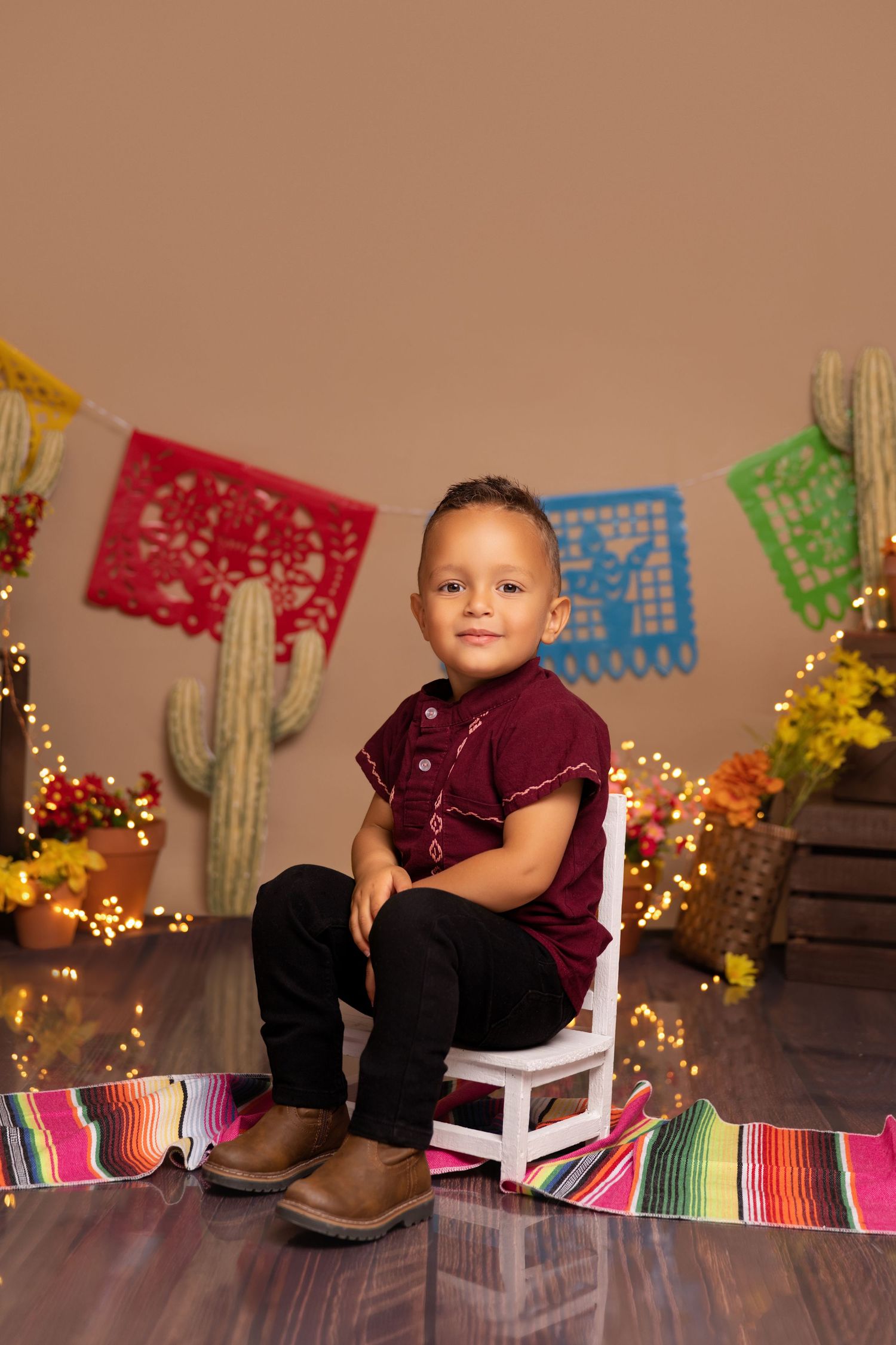 Young boy in maroon shirt sits on white chair with colorful papel picado banners and cacti in festive Mexican-themed portrait.