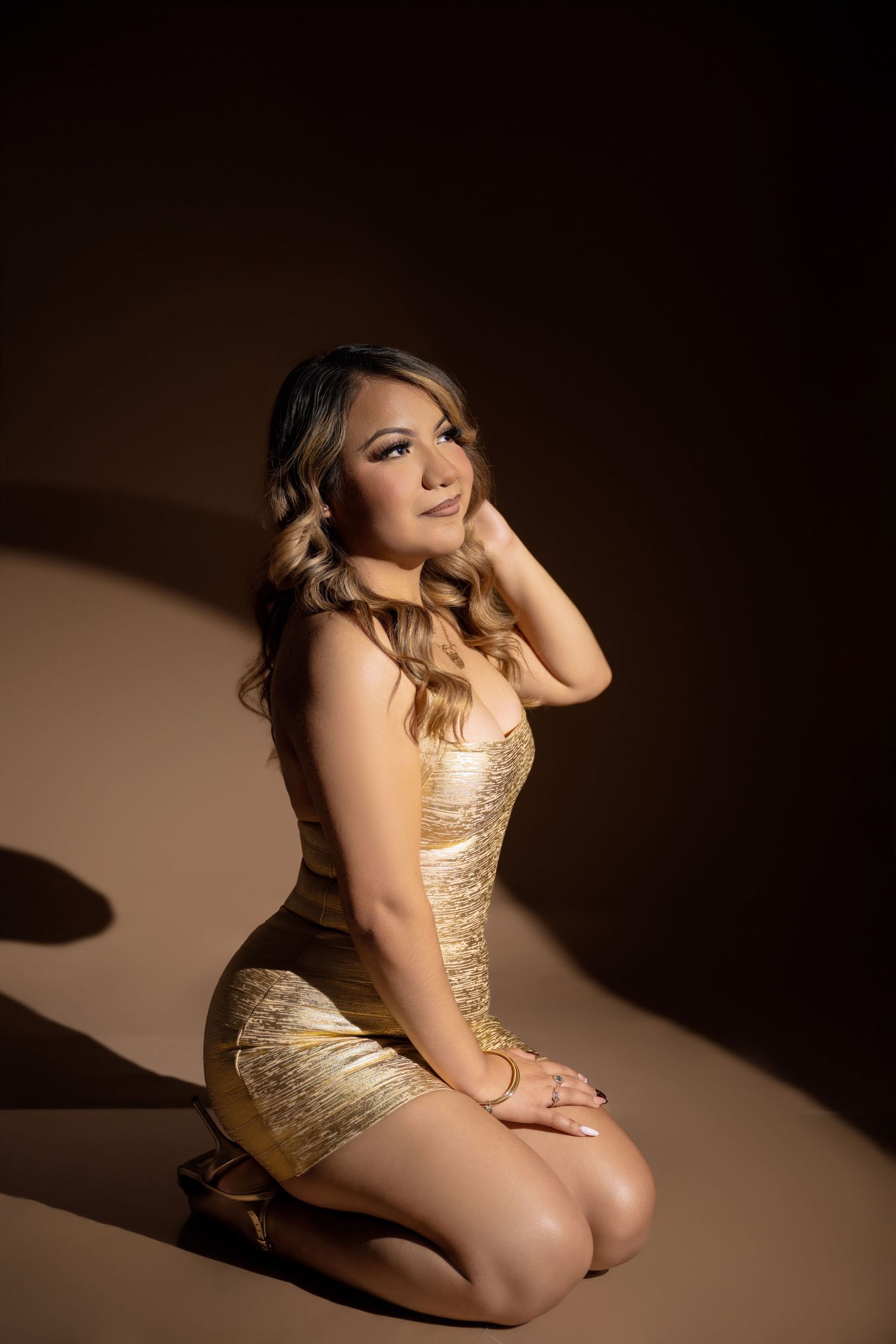 Woman with long wavy hair kneeling in a gold metallic bodycon dress and heels against a dark brown studio backdrop with dramatic lighting.
