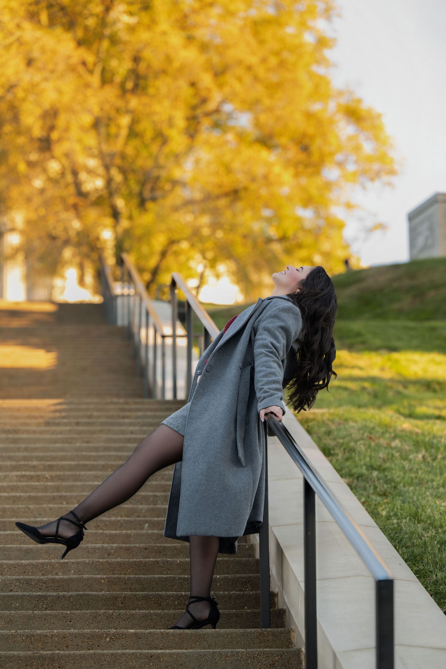 Woman in a gray coat and black heels leans back on a stone staircase railing beneath golden autumn trees.