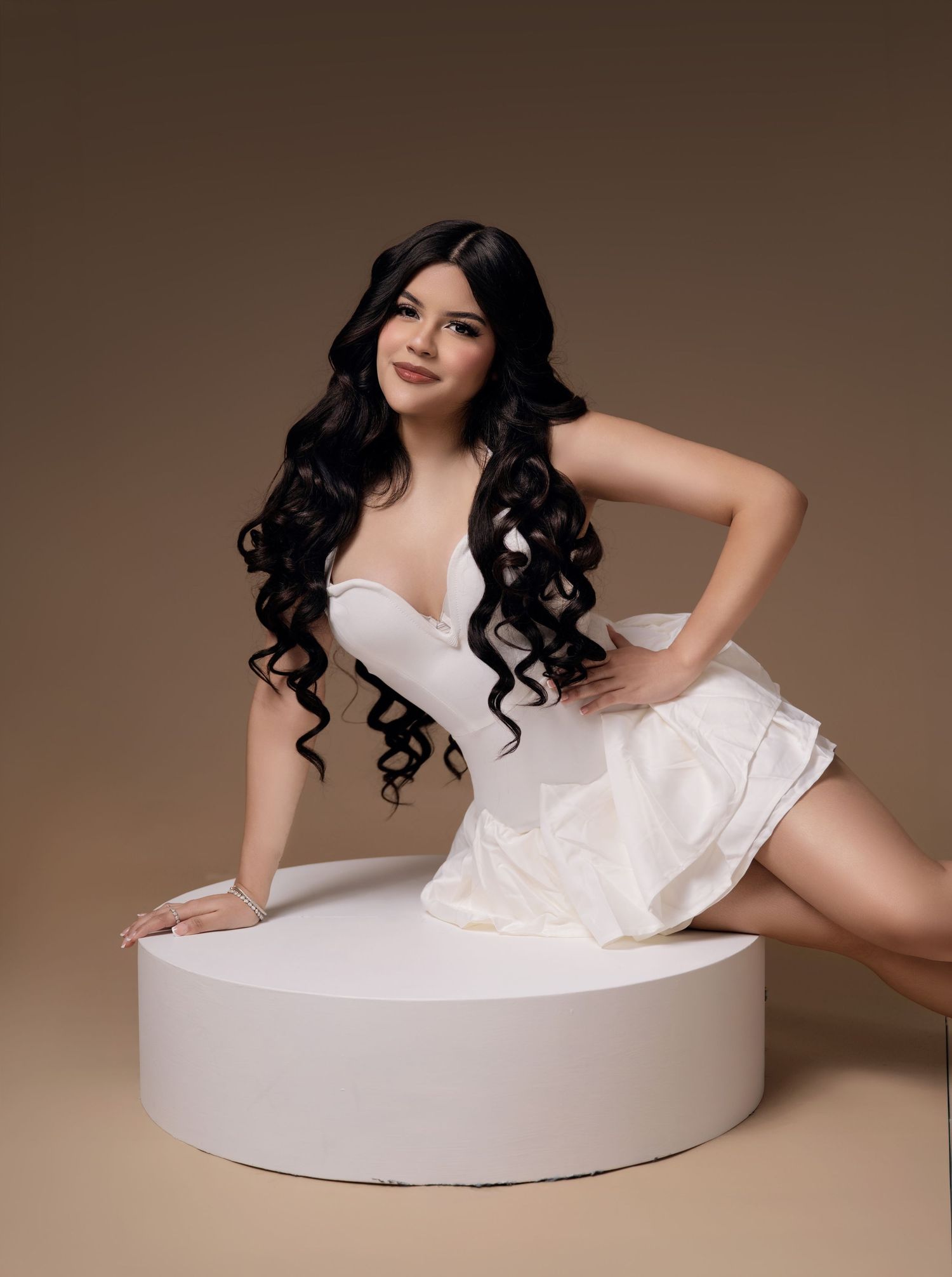 Woman with long black curly hair in a white strapless mini dress posing on a white round platform against a beige studio backdrop.