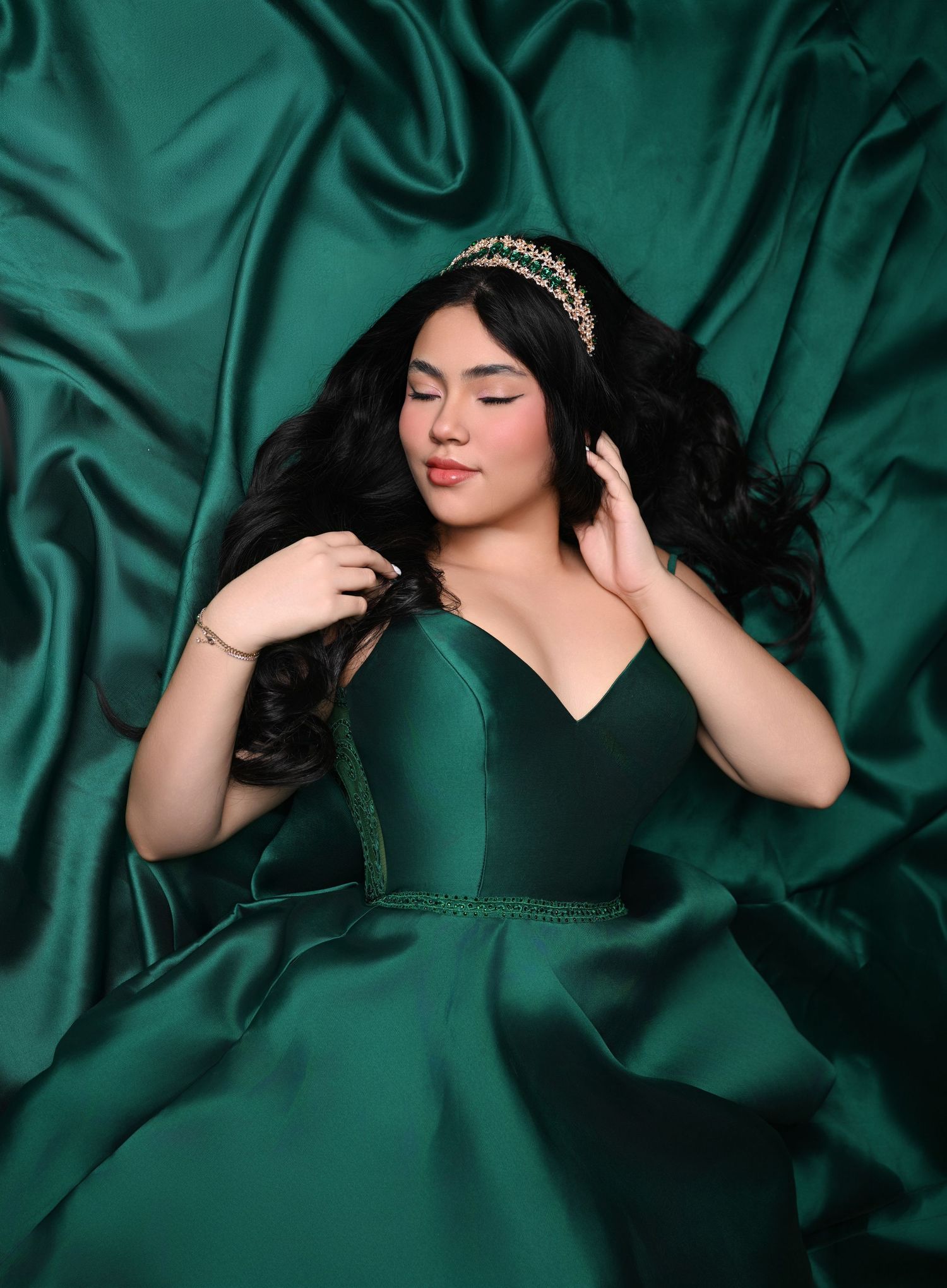 Young woman in emerald green satin gown and gold tiara poses gracefully against lush green satin fabric backdrop.