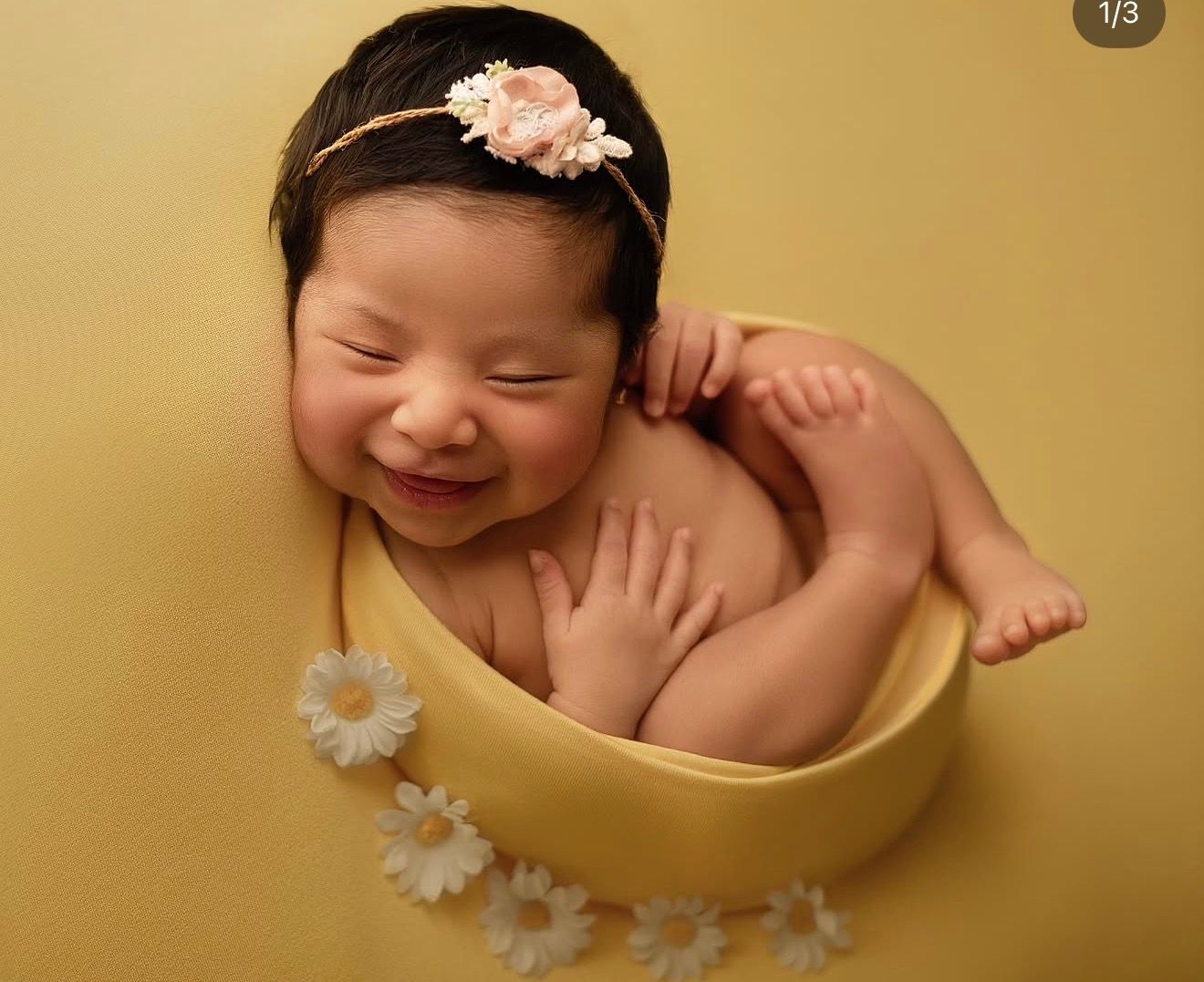 Smiling newborn baby girl in yellow bowl with daisy flowers, wearing pink floral headband, professional newborn photography.