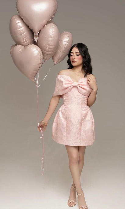 Woman in a pink bow-detail mini dress and strappy heels holds a bunch of blush heart-shaped balloons against a neutral studio backdrop.