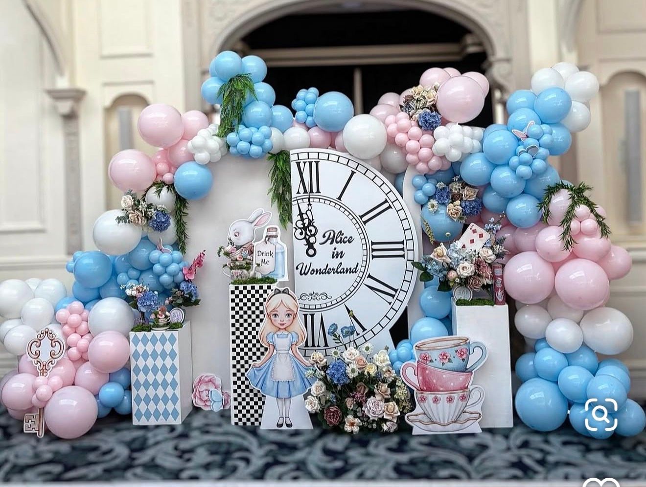 Alice in Wonderland party backdrop with pink and blue balloon arch, giant clock, and character decorations.