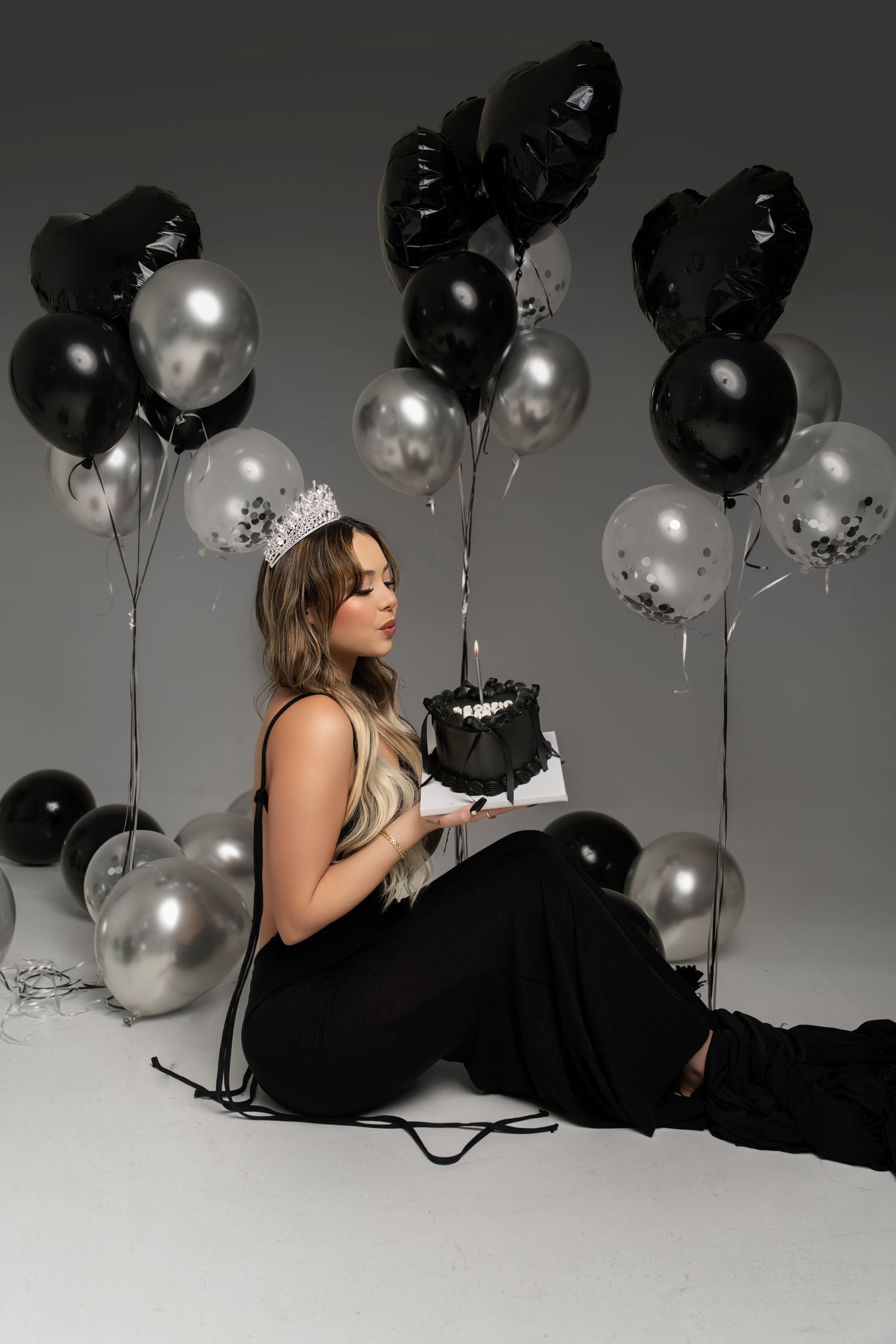 Elegant party scene with black and silver balloons surrounding someone in black dress holding a small dark cake.
