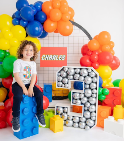 Colorful balloon arch and large LEGO-inspired building blocks create a vibrant birthday party backdrop with playful elements.