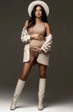 Model in neutral tones poses in white boots and blazer against dark background for fashion photoshoot.