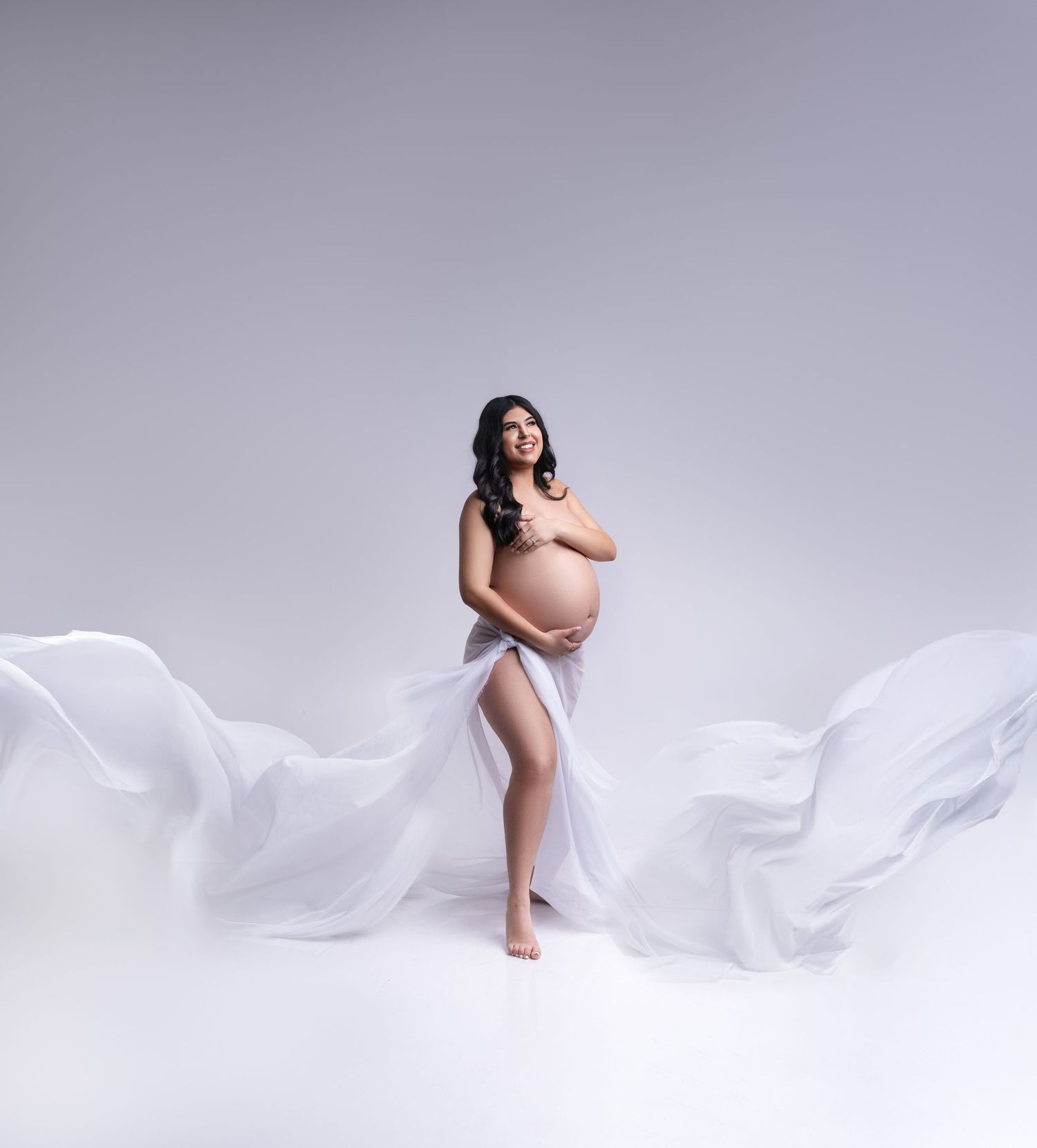 Ethereal maternity photo with flowing white fabric against a light gray background creating an artistic silhouette.