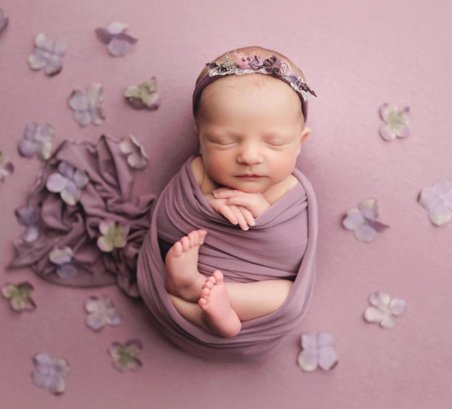 Newborn baby wrapped in purple fabric lying on pink backdrop with scattered paper butterflies around them.