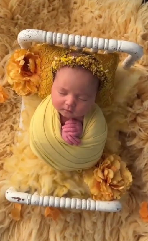 A sleeping newborn wrapped in soft yellow fabric lies on a fuzzy blanket with yellow flowers and white wooden props.