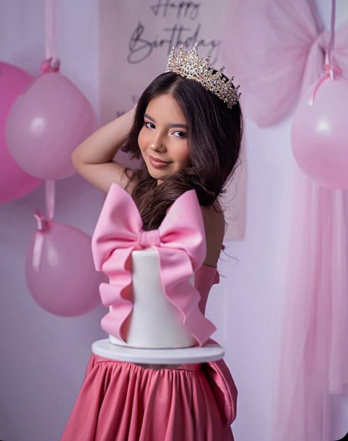 Young girl in pink bow dress and tiara poses with white birthday cake against pink balloon backdrop.