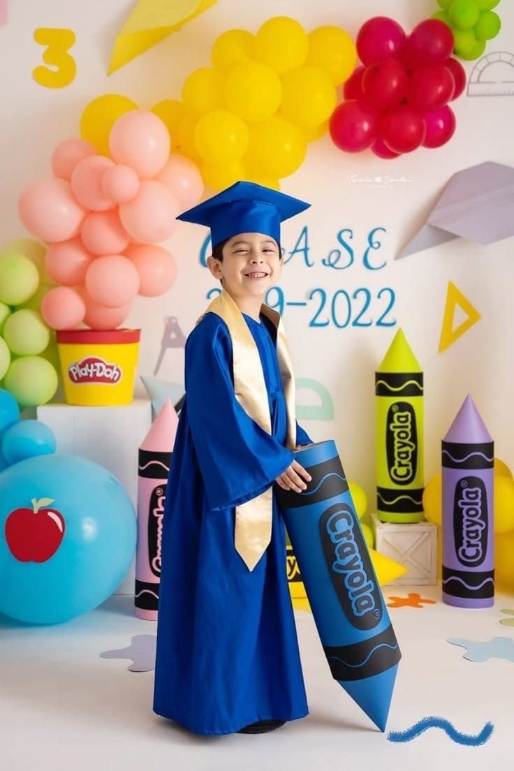 Child in blue graduation cap and gown holding giant Crayola crayon at colorful school-themed photo backdrop.
