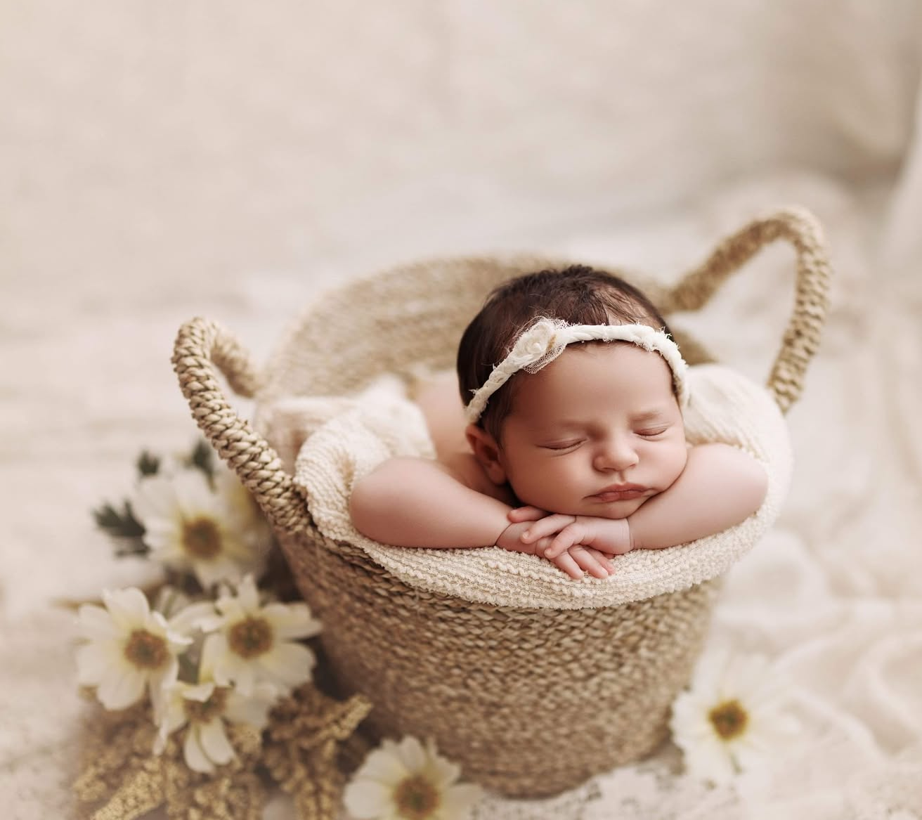 Sleeping newborn baby in a woven basket with white daisy flowers and lace headband on cream background.