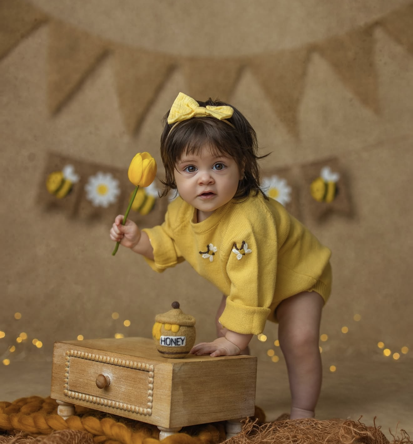 Baby girl in yellow outfit holds tulip near honey pot, bee-themed birthday photoshoot with bunting backdrop.