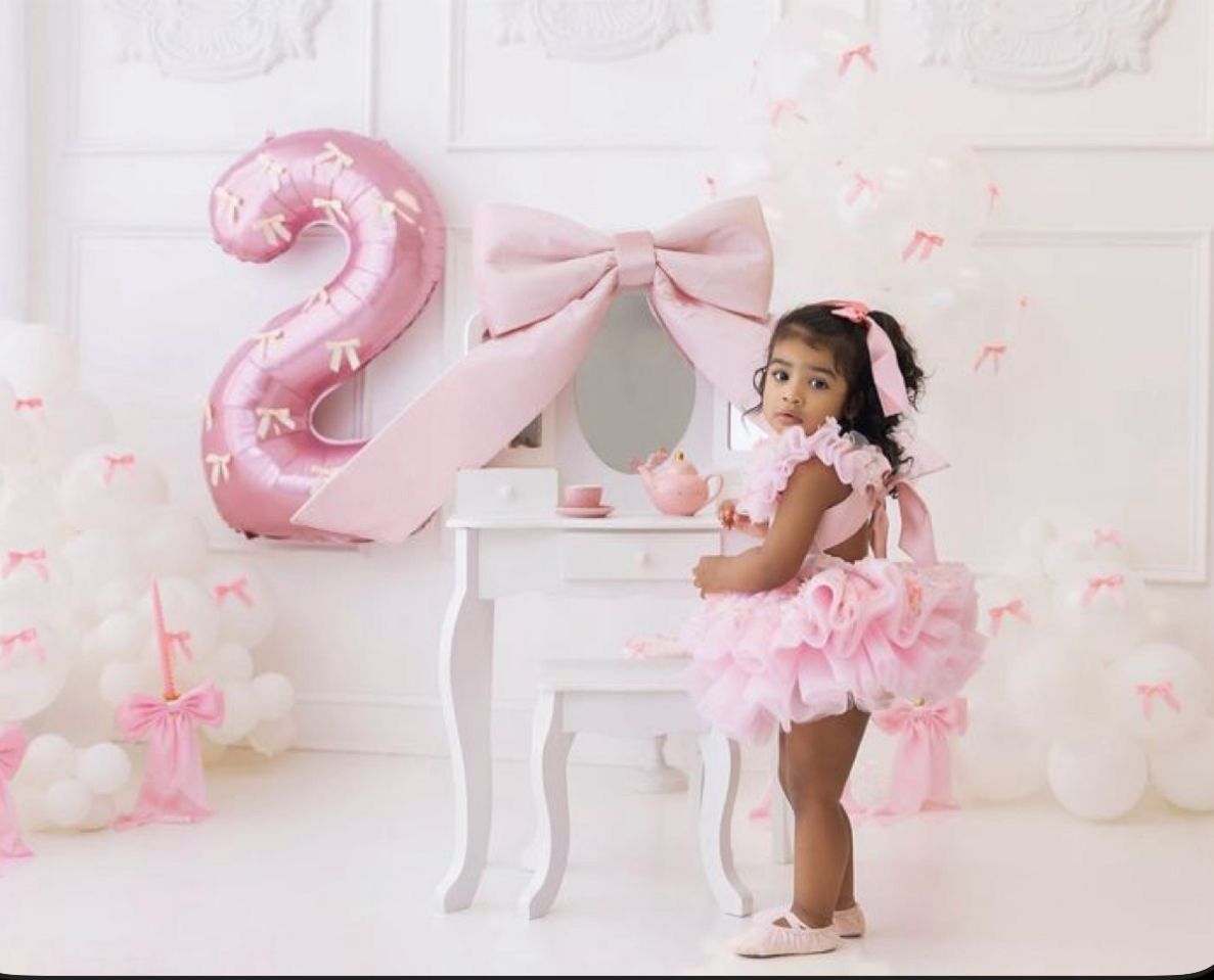 Toddler girl in pink tutu dress poses by white vanity table with large pink number 2 balloon at birthday party.