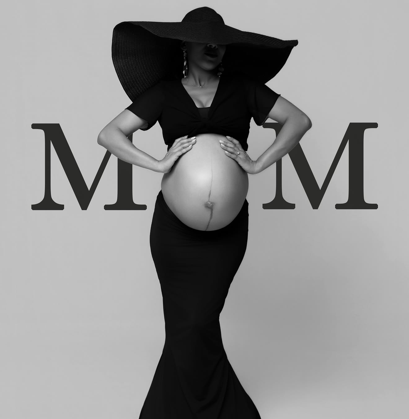 Black and white maternity photo of pregnant woman in black gown and wide-brim hat posing with MOM text.