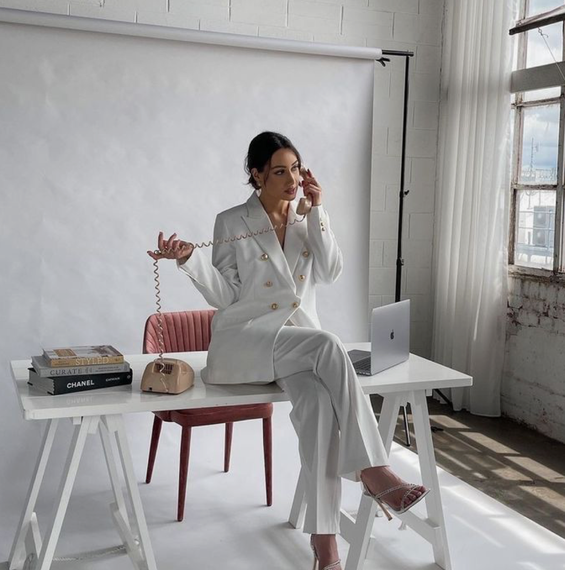 Woman in white blazer suit sits on desk with Chanel bag, laptop, and books in bright studio setting.