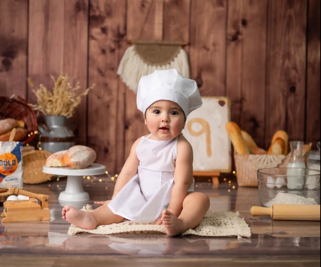 A small baby in a chef's hat and apron sits on a wooden counter surrounded by baking ingredients and supplies.