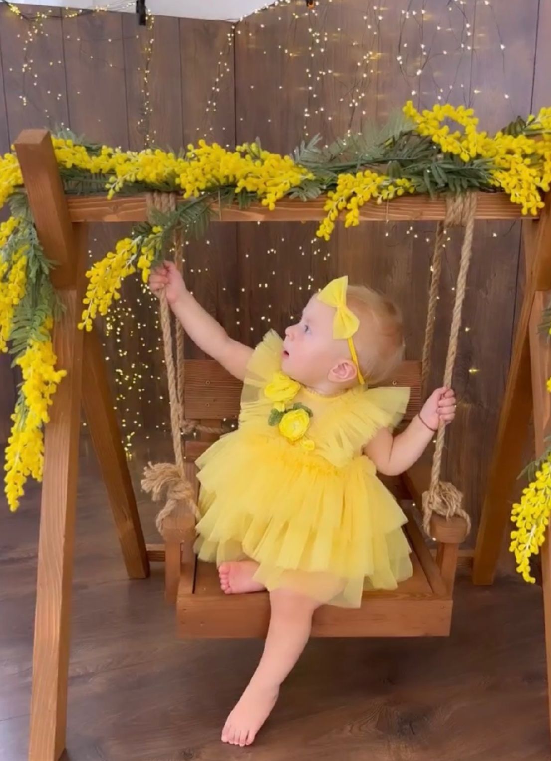 A baby in a yellow tutu dress poses beneath a wooden archway decorated with twinkling lights and yellow flowers.