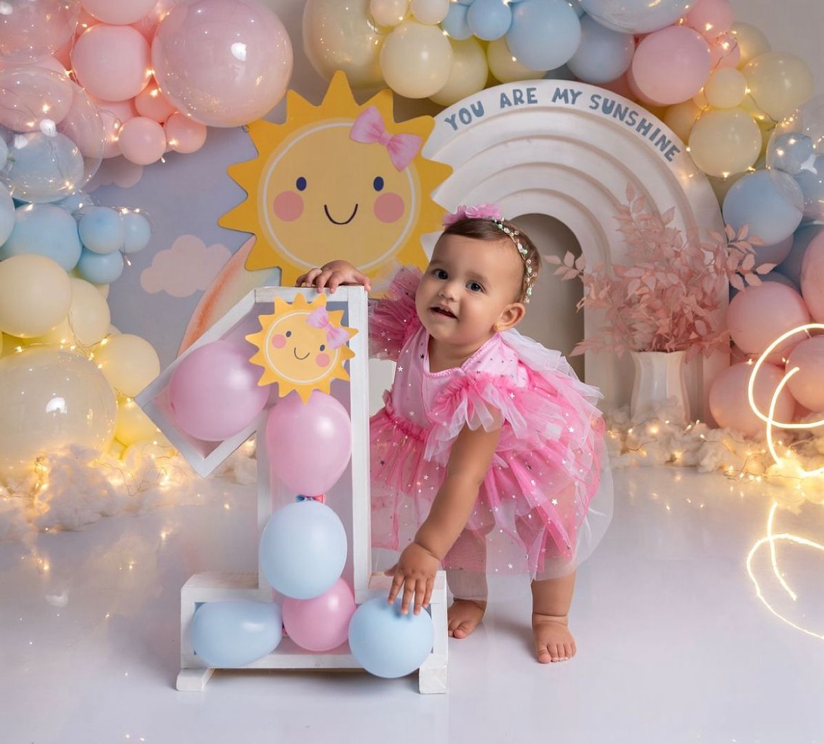 Pastel balloon arch with pink and blue hues decorated with string lights and a smiling sun cutout creates a dreamy backdrop.