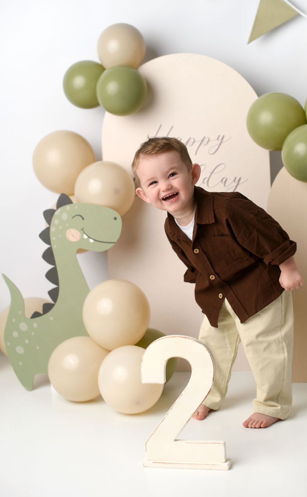A child in brown sweater poses playfully against a backdrop of green and cream colored balloons and dinosaur cutout decorations.