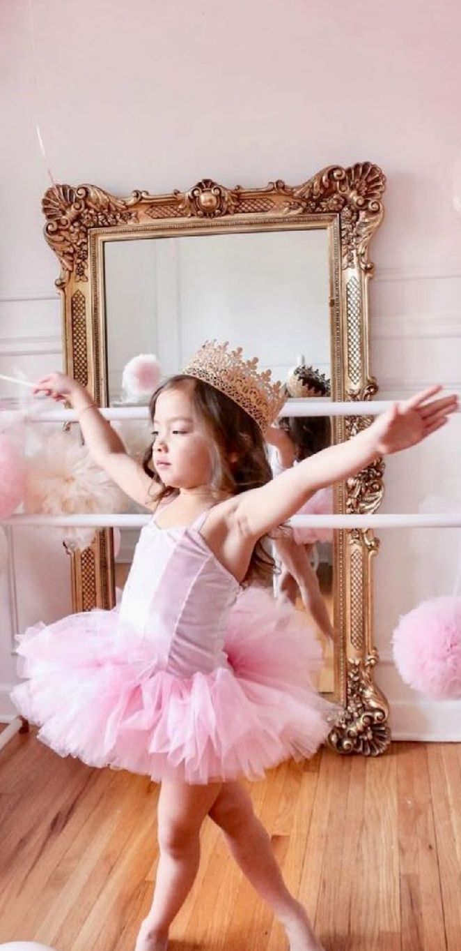 A dancer in a pink tutu performs graceful ballet poses in front of an ornate gold-framed mirror on hardwood floors.
