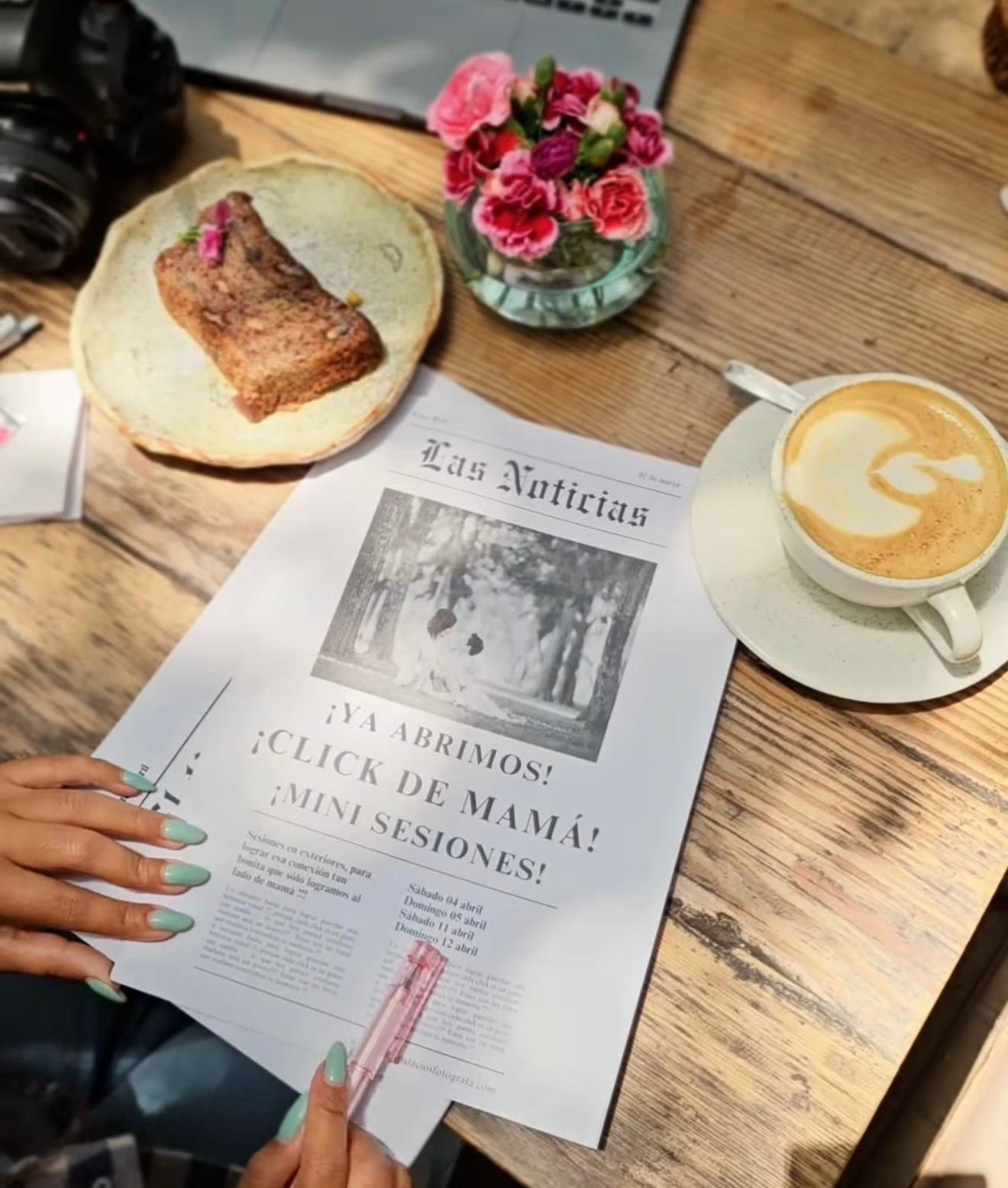 A latte and pastry beside a Spanish newspaper Las Noticias on a wooden cafe table with pink roses.