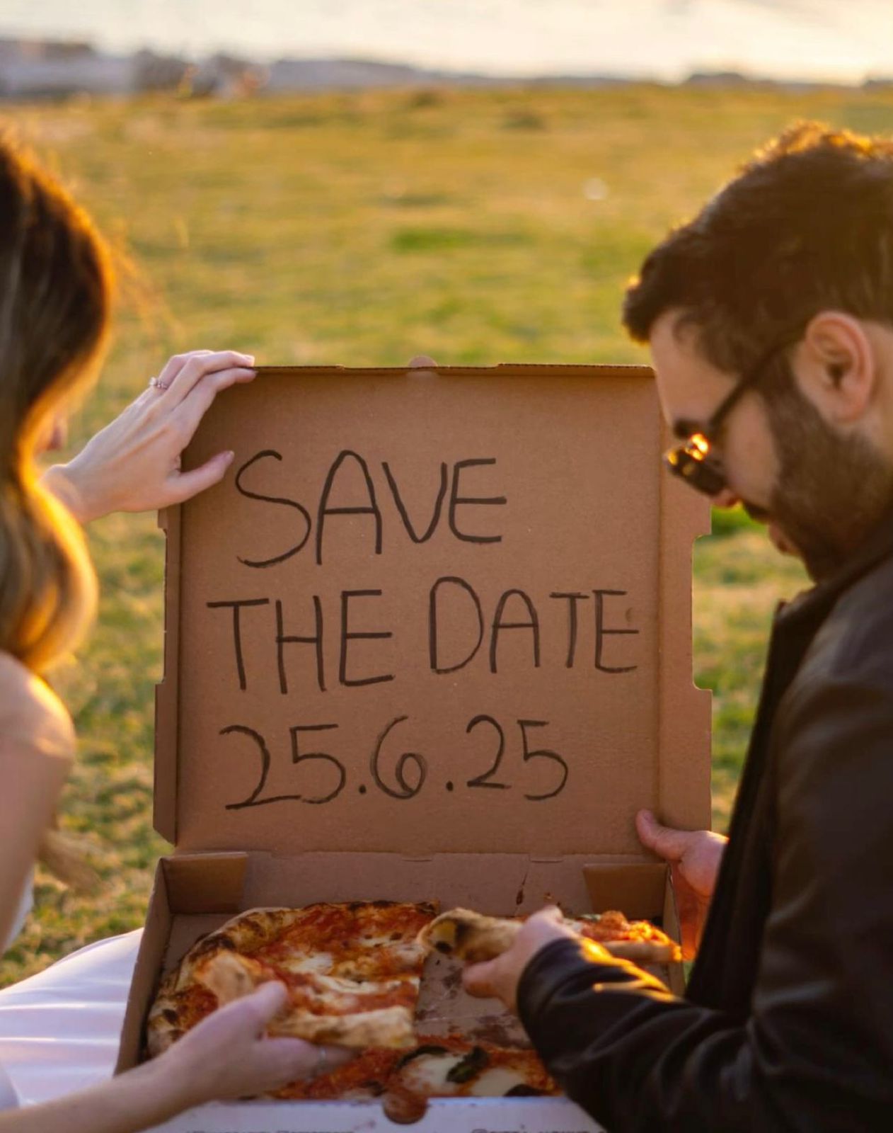Couple holding a pizza box with Save the Date 25.6.25 written on it outdoors at golden hour.