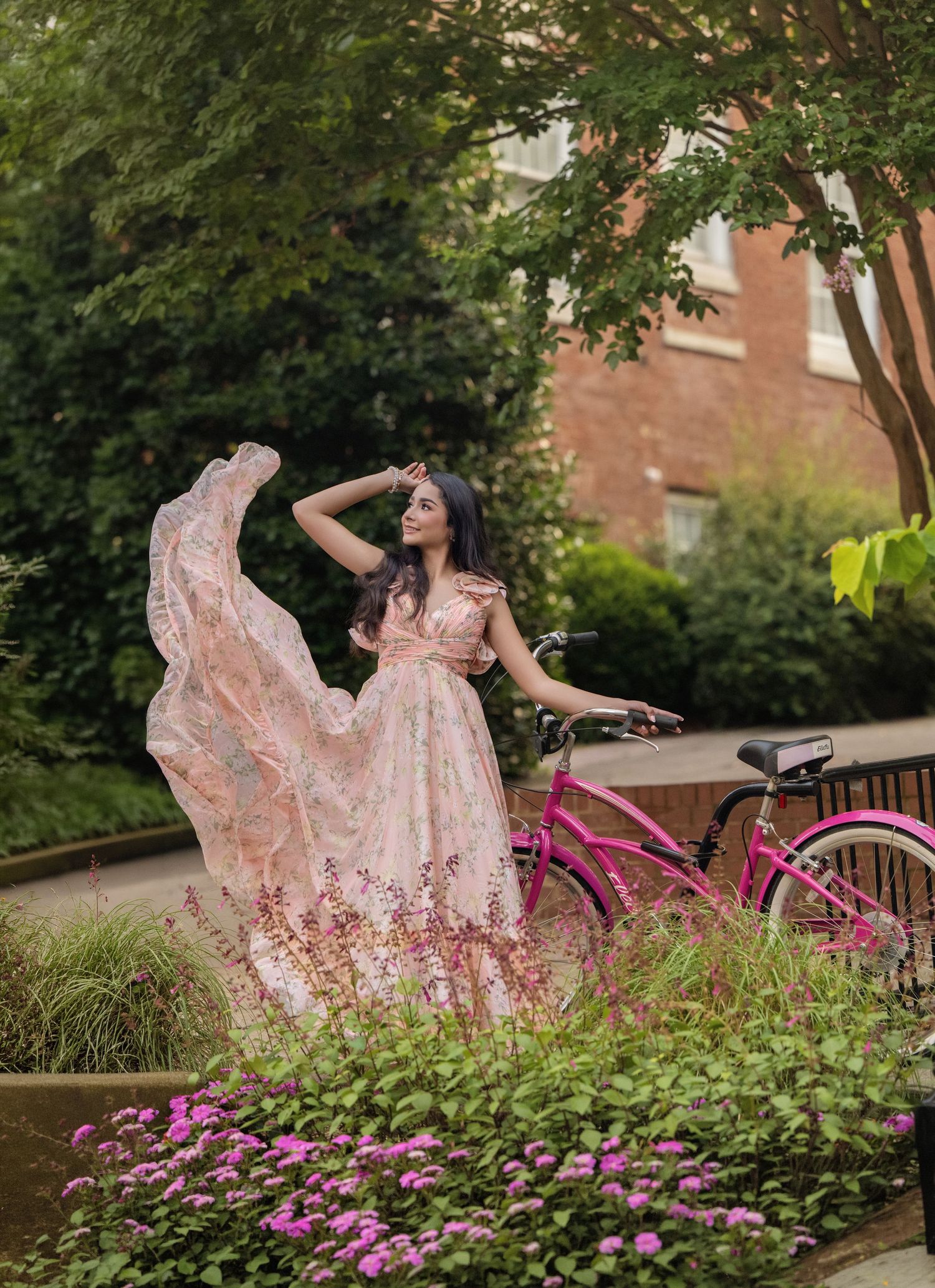 Woman in flowing pink floral gown poses gracefully near a pink bicycle surrounded by lush greenery and flowers.