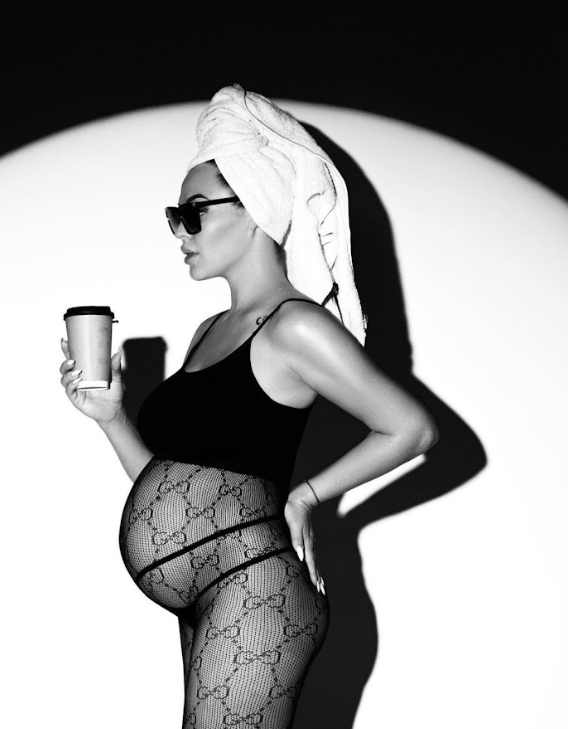 Black and white photo of a pregnant figure in designer patterned tights and head wrap holding a coffee cup.