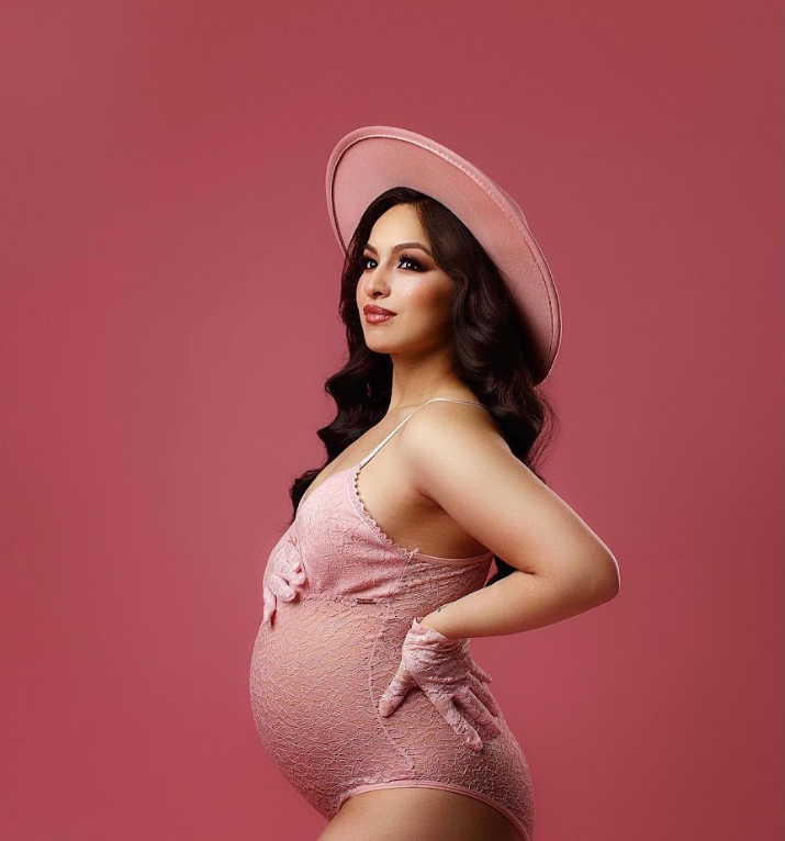Vintage-inspired maternity portrait with pink hat and dress against rose-colored backdrop.