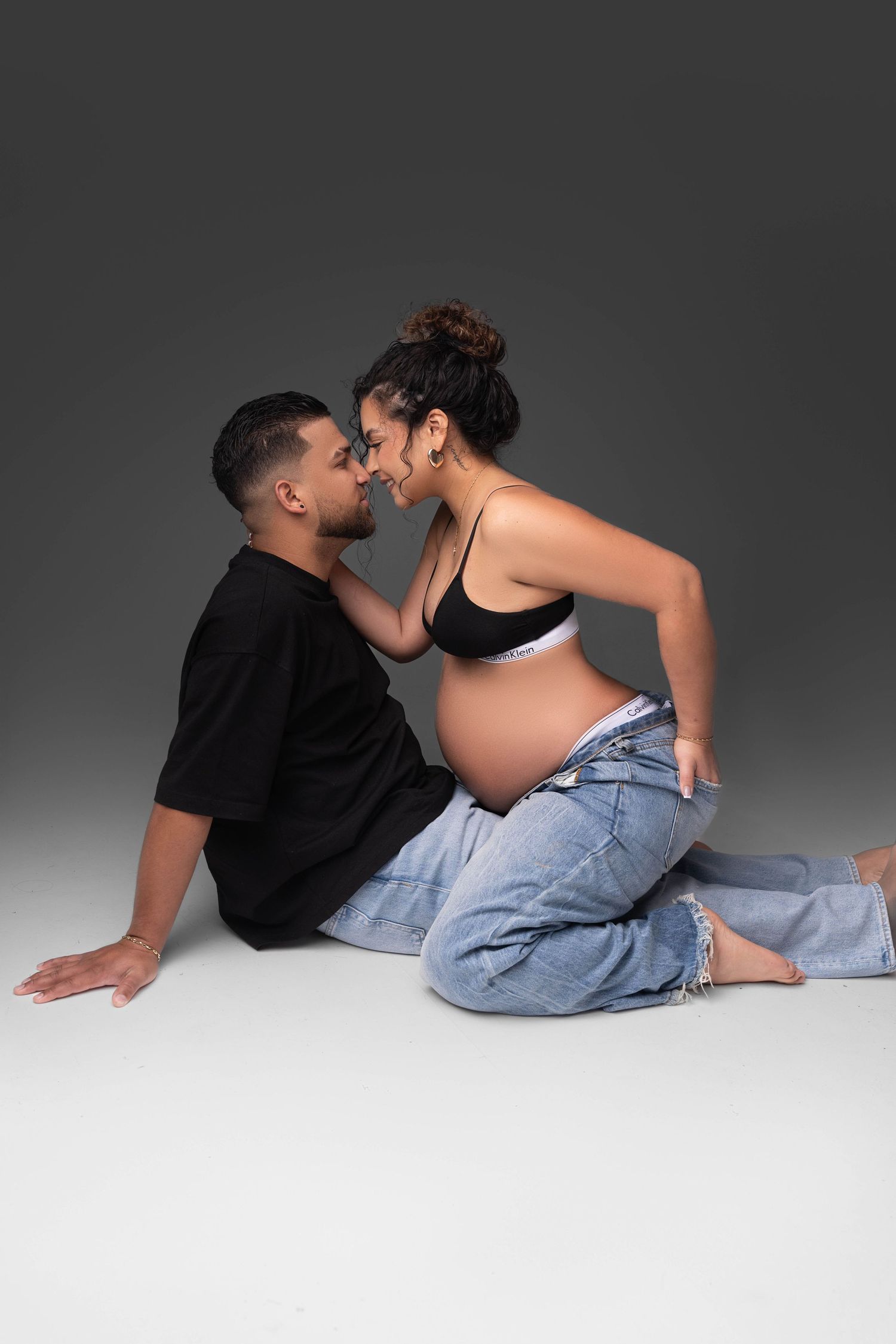 A couple in casual attire embraces intimately while lying on a light gray floor in an artistic photo series.