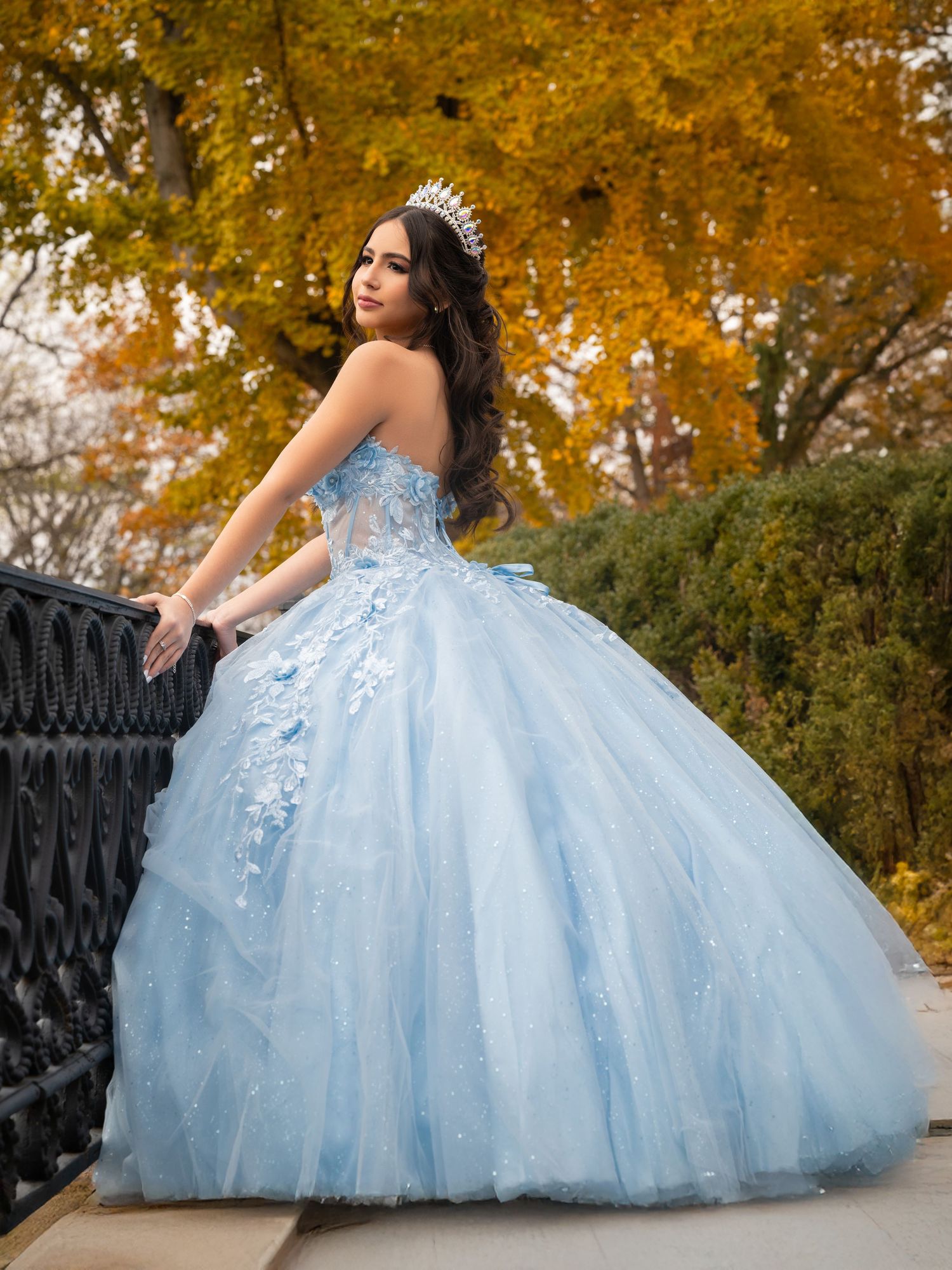 Young woman in a light blue ball gown and tiara poses gracefully outdoors with autumn foliage backdrop.