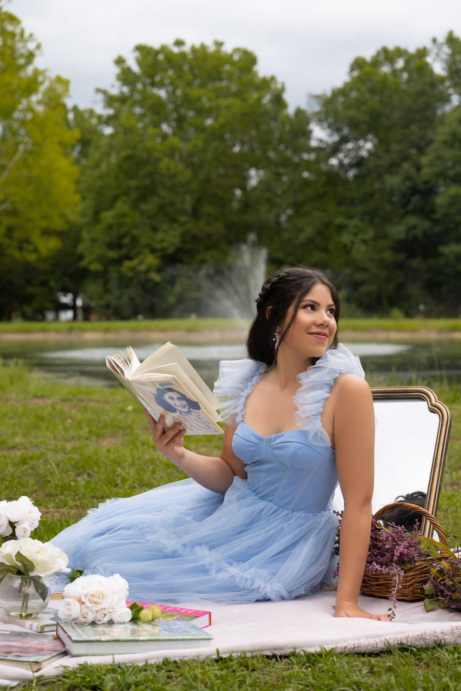 Young woman in blue tulle dress reads a book at a park picnic near a fountain, surrounded by flowers and greenery.