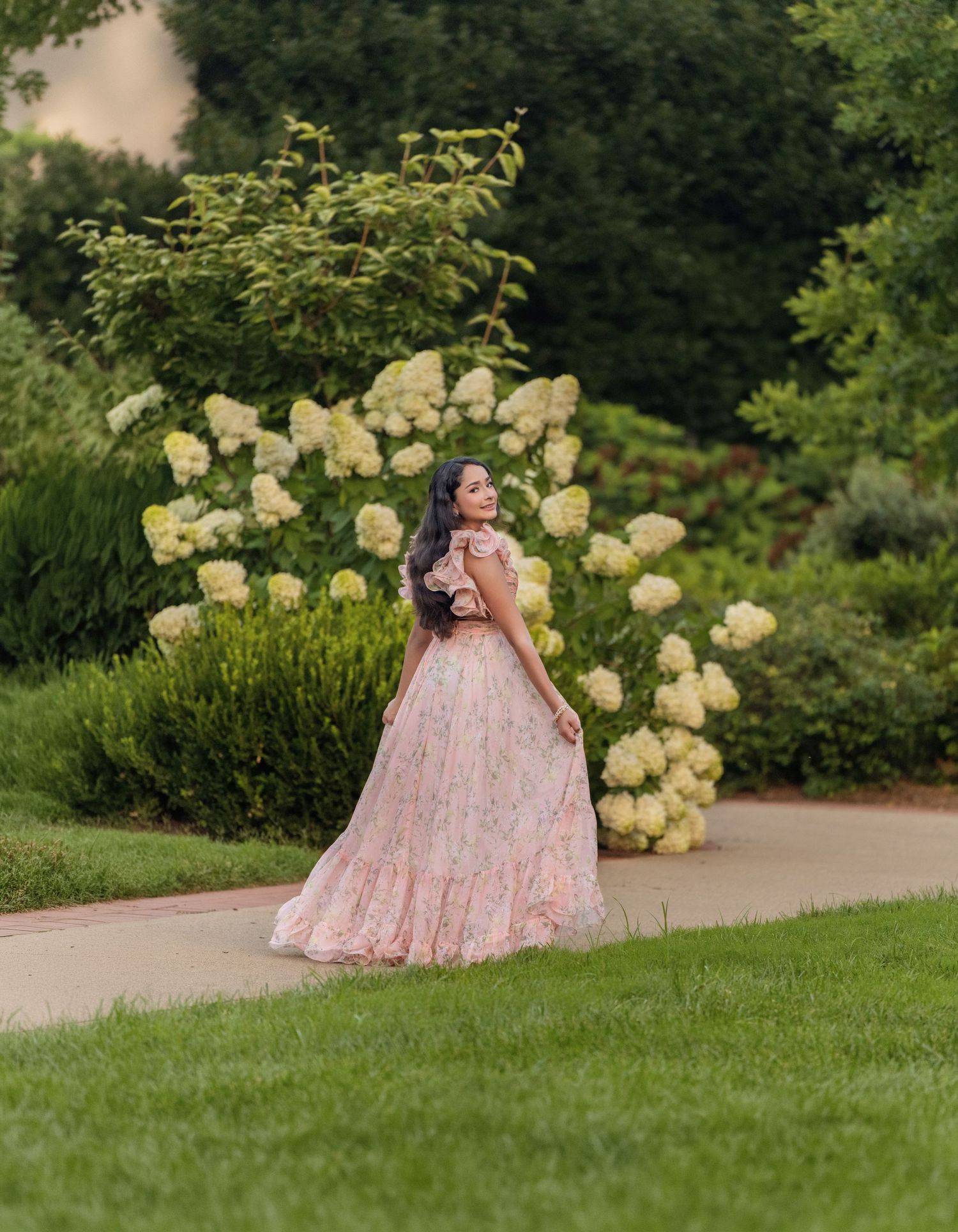 Woman in pink floral off-shoulder lehenga twirling on garden path with white hydrangeas blooming behind her.
