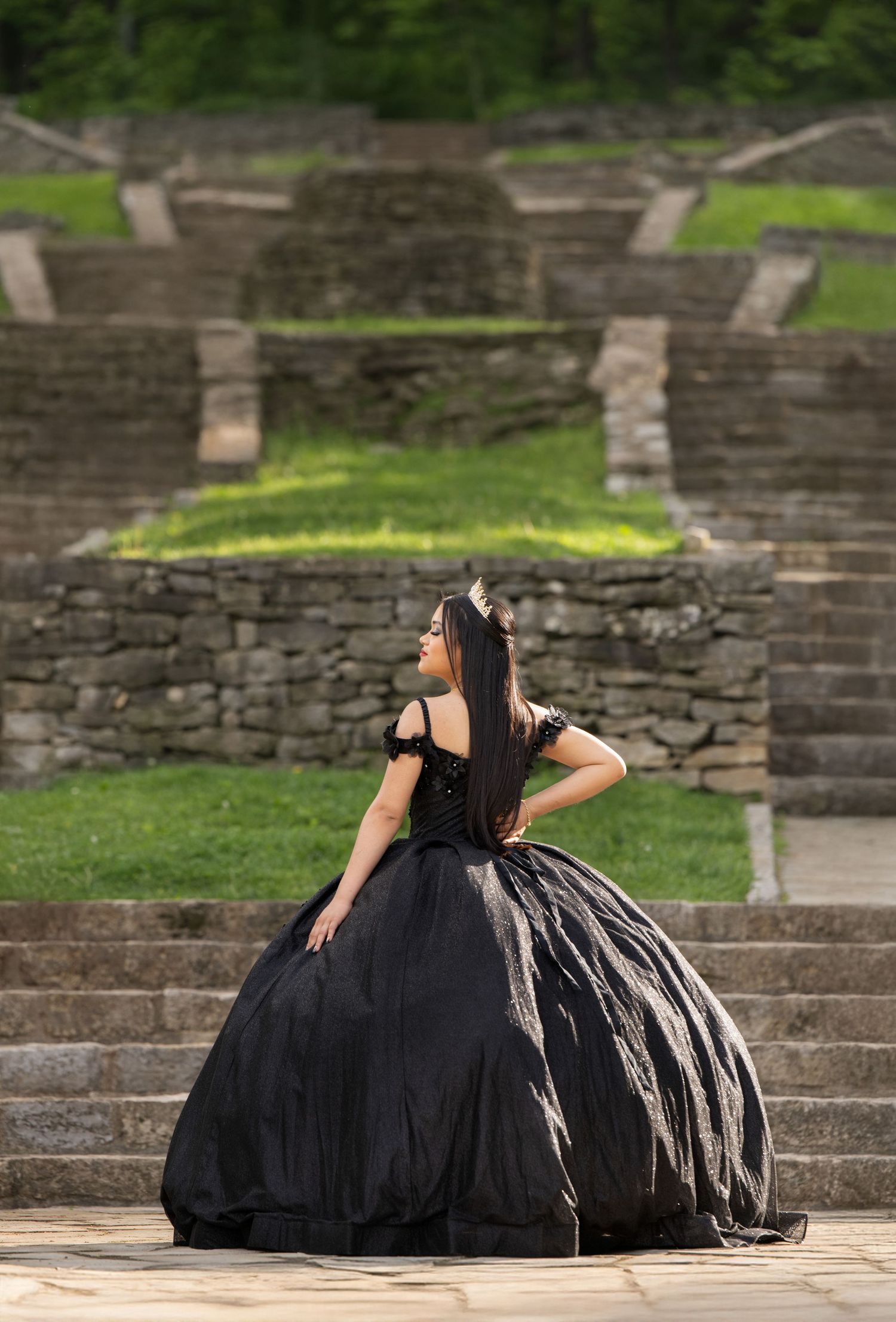 Young woman in elegant black ball gown poses on stone steps at an outdoor amphitheater surrounded by lush greenery.