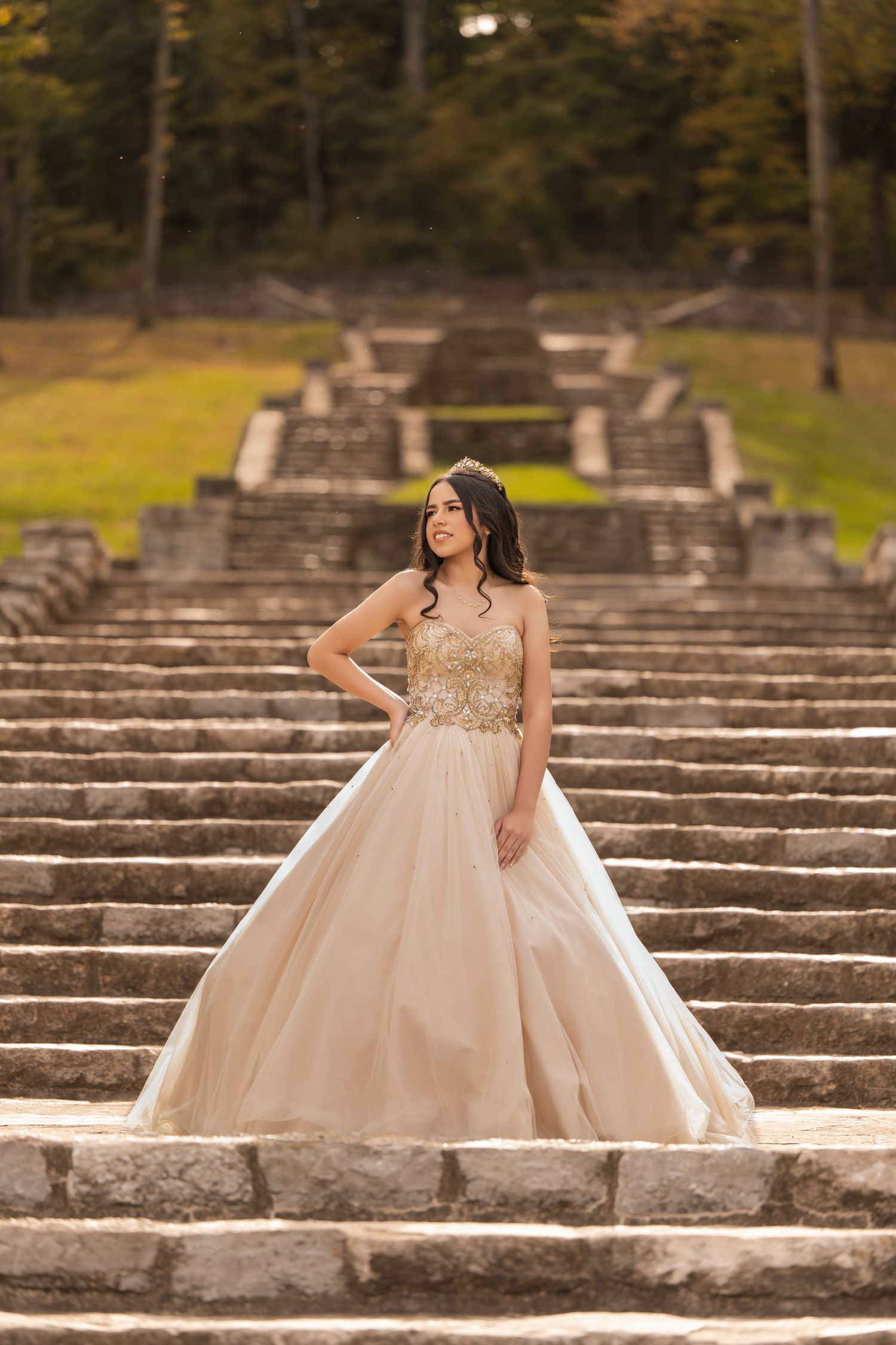 Young woman in a champagne ballgown with gold embroidery and tiara poses on stone steps outdoors.