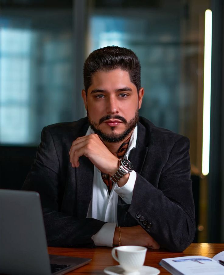 Professional businessman in dark suit sitting at desk with laptop, coffee cup, and documents in modern office.