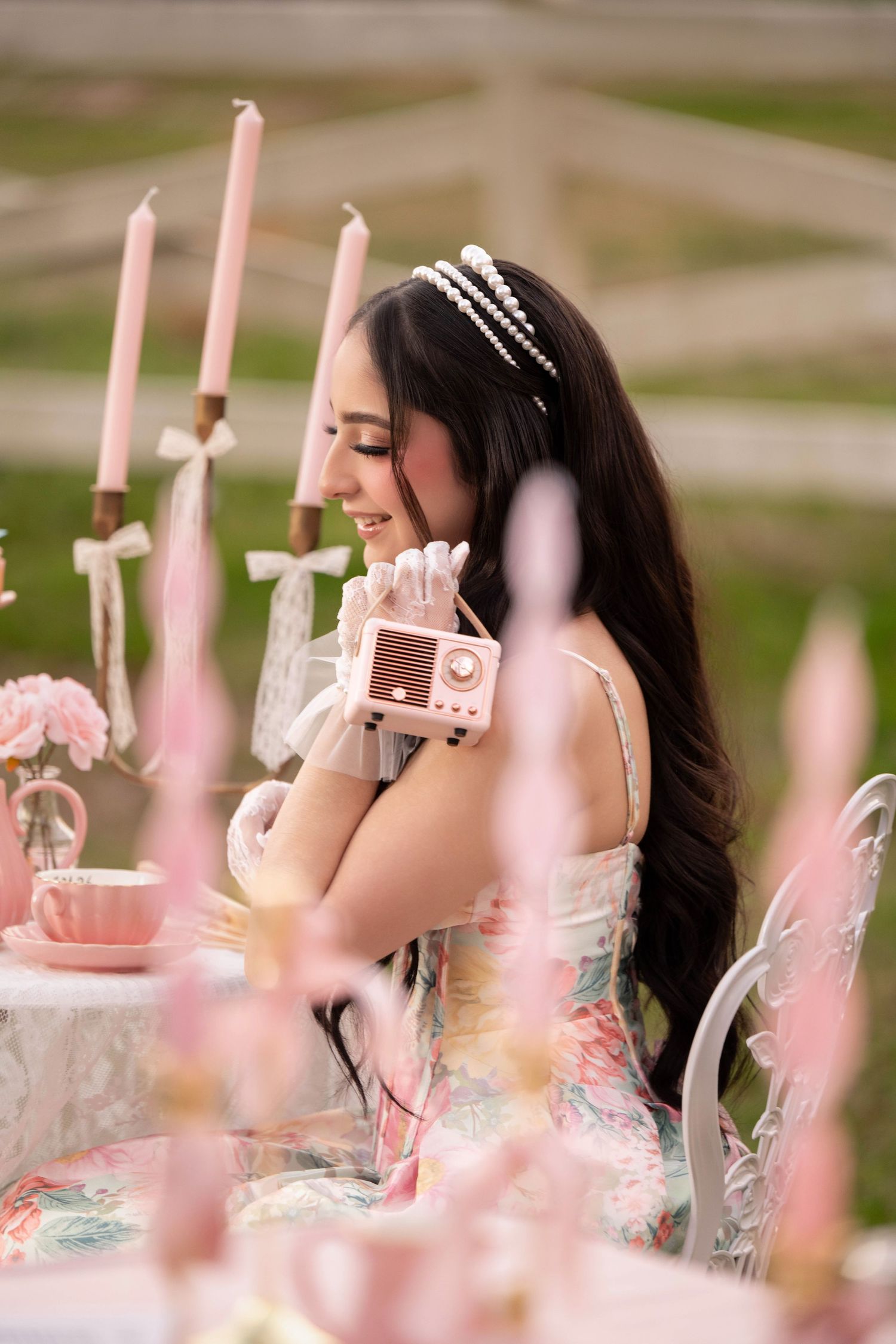 Young woman in floral dress holding pink vintage radio at elegant outdoor tea party with pink candles and roses.