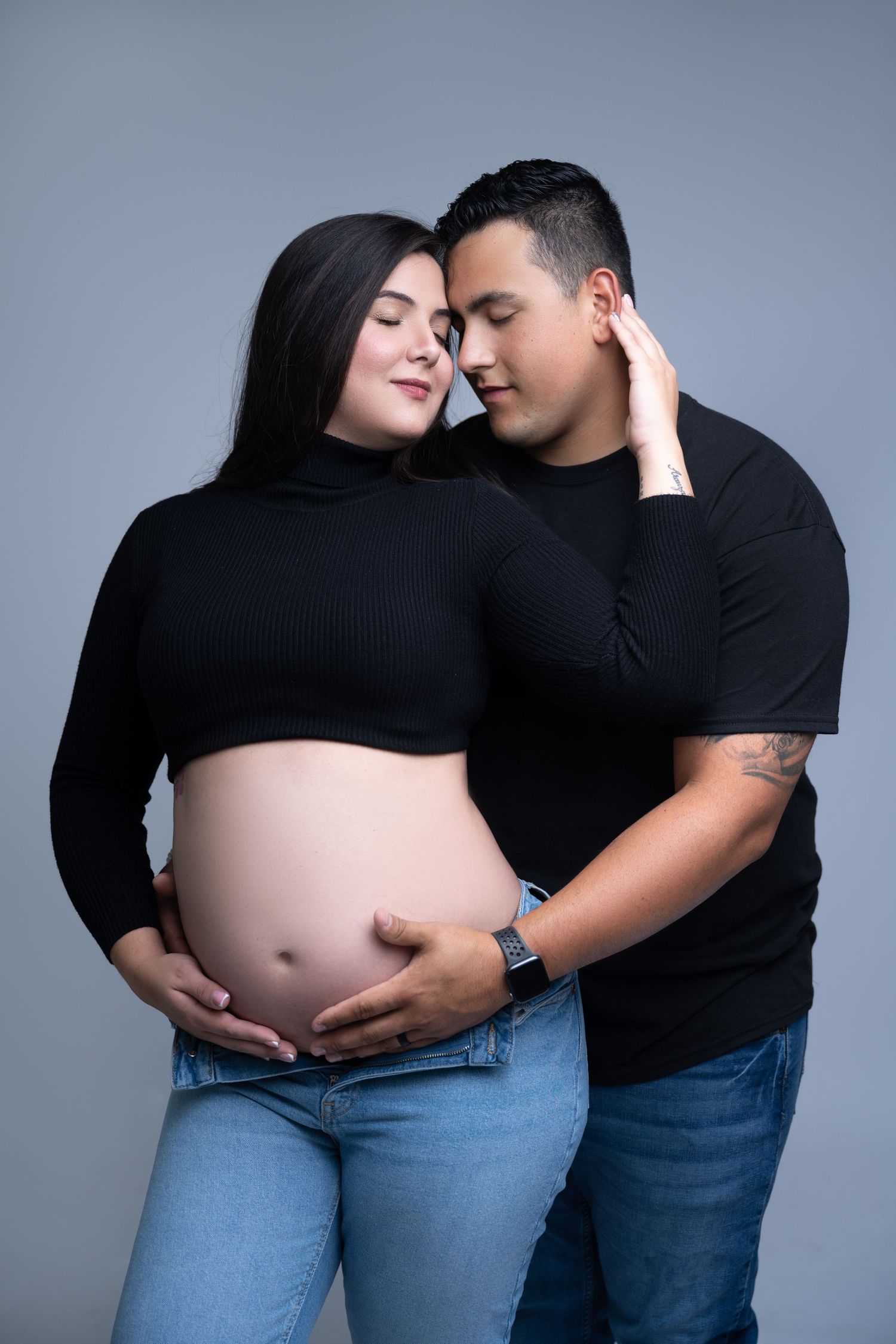 Expectant couple in black shirts embrace tenderly against gray background during maternity photoshoot.