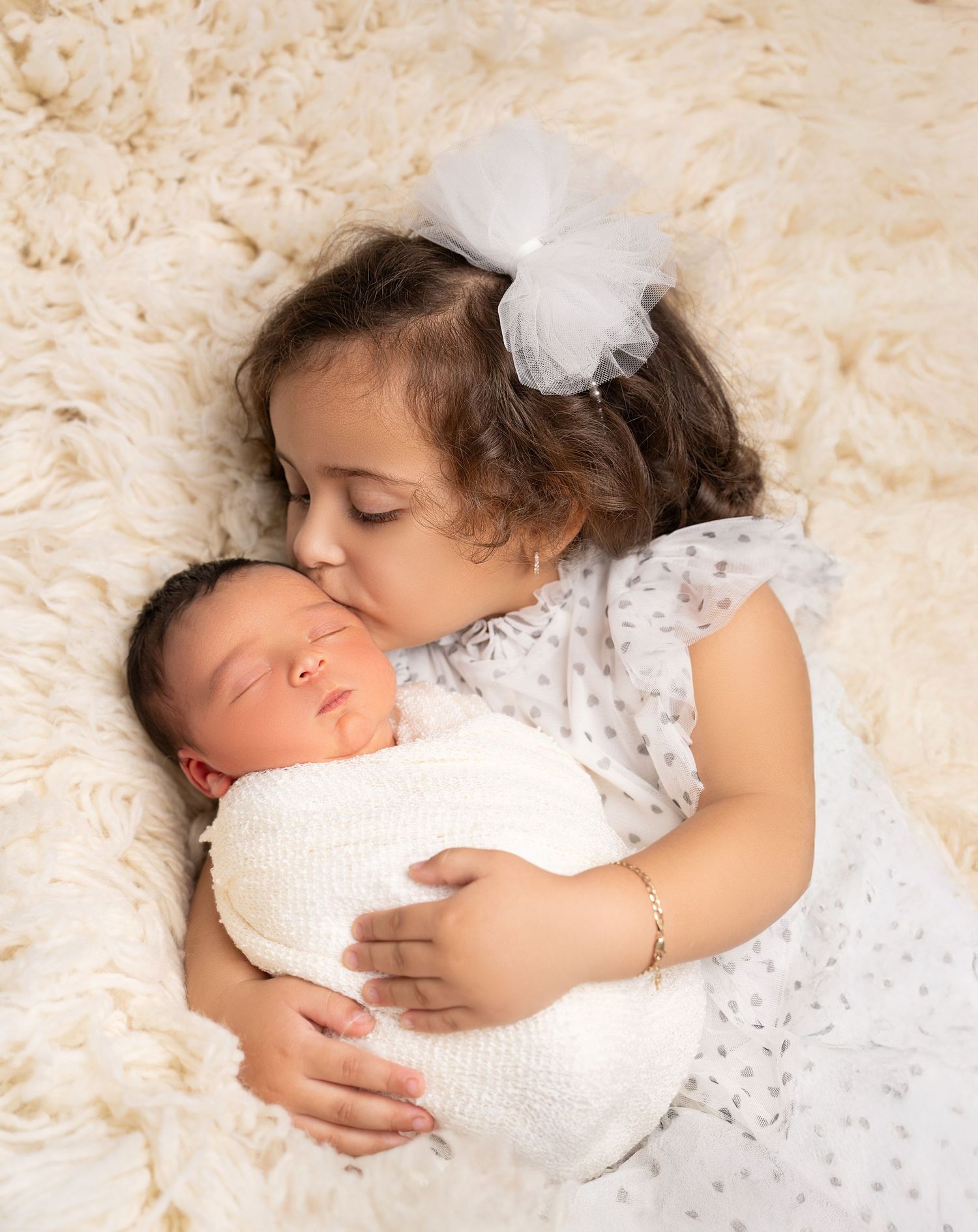 Tender moment between siblings on cream fluffy rug with newborn wrapped in white.
