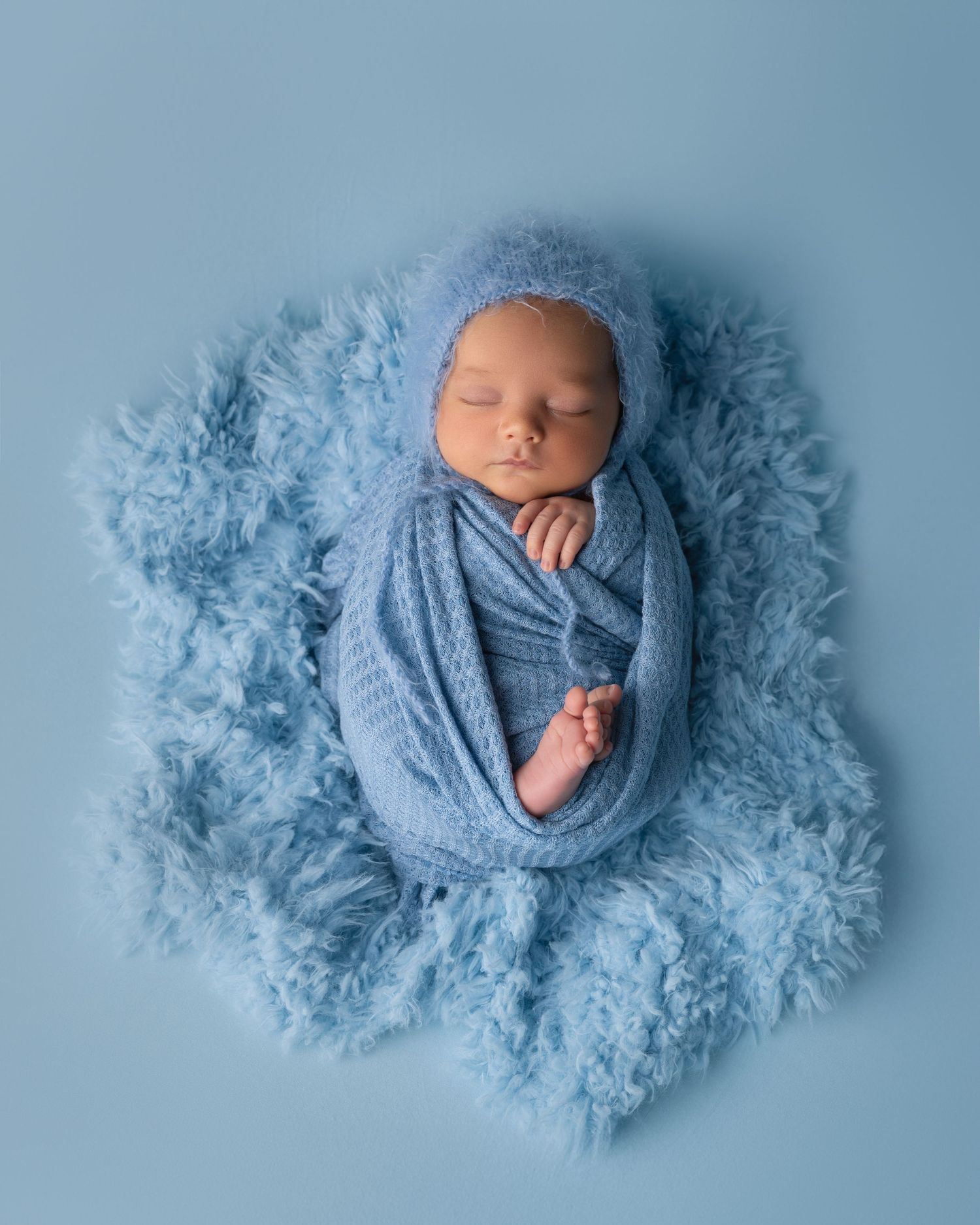 Peaceful sleeping newborn wrapped in blue knit on matching fluffy backdrop.