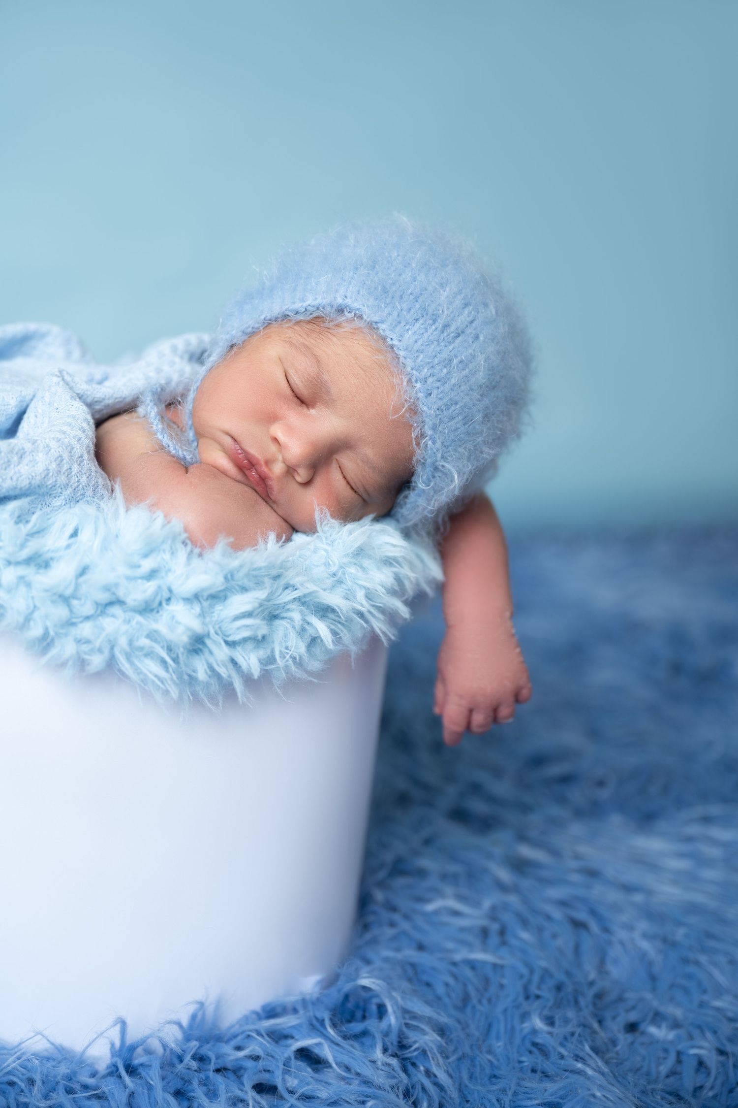 Sleeping newborn baby in blue knit hat resting in white bucket with blue fur lining on blue rug.