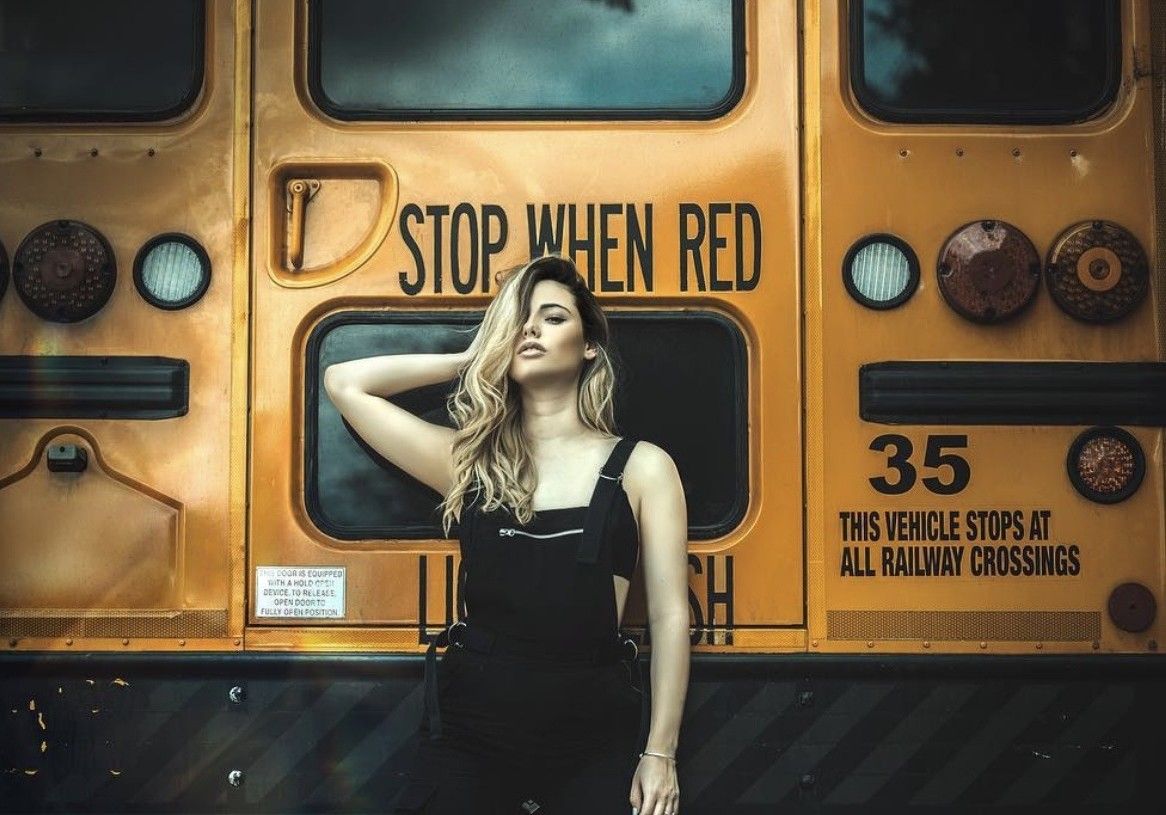 Young blonde woman in black overalls poses in front of a yellow school bus with Stop When Red sign.